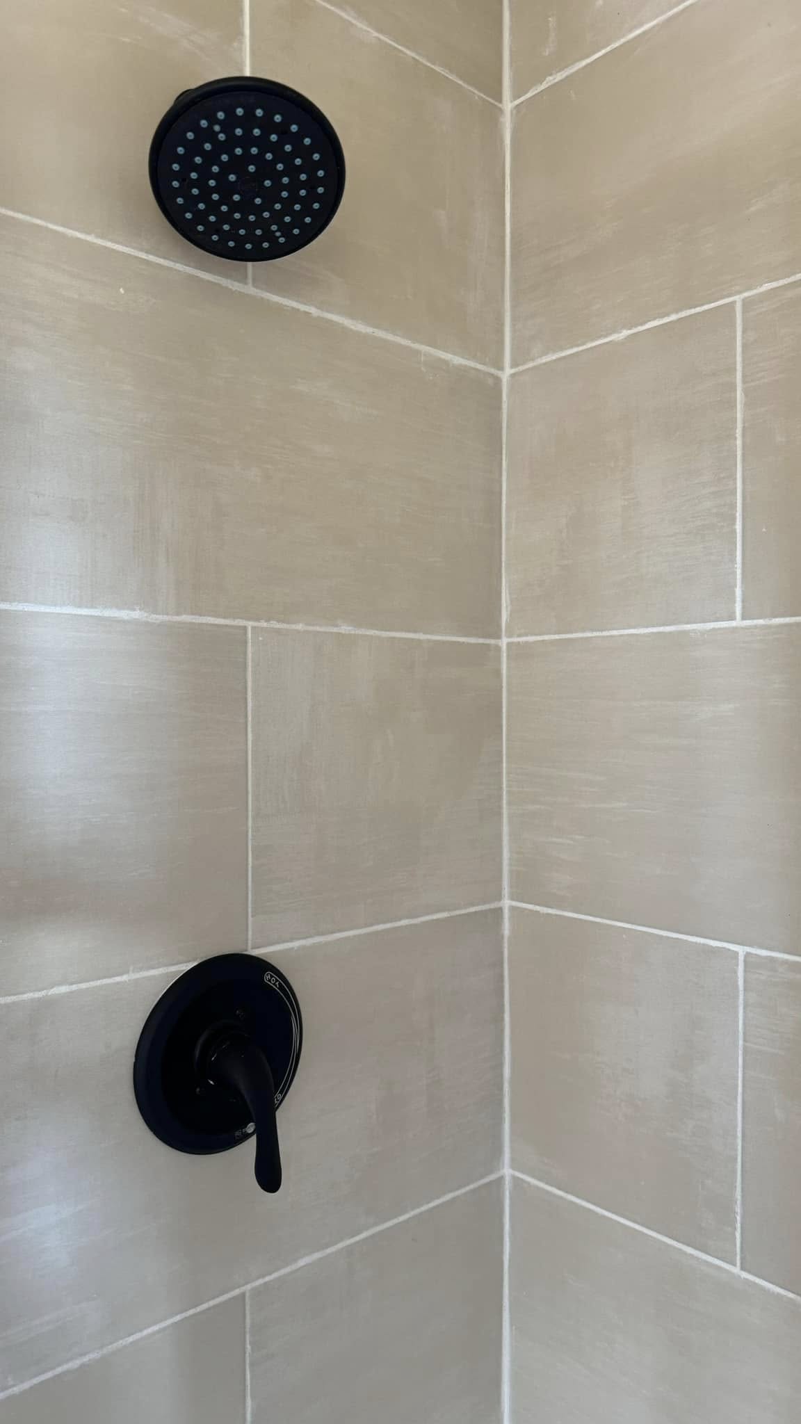 Alt text: A shower with black fixtures, featuring a round showerhead and a single lever handle against beige tiles. The scene is clean and minimalistic.