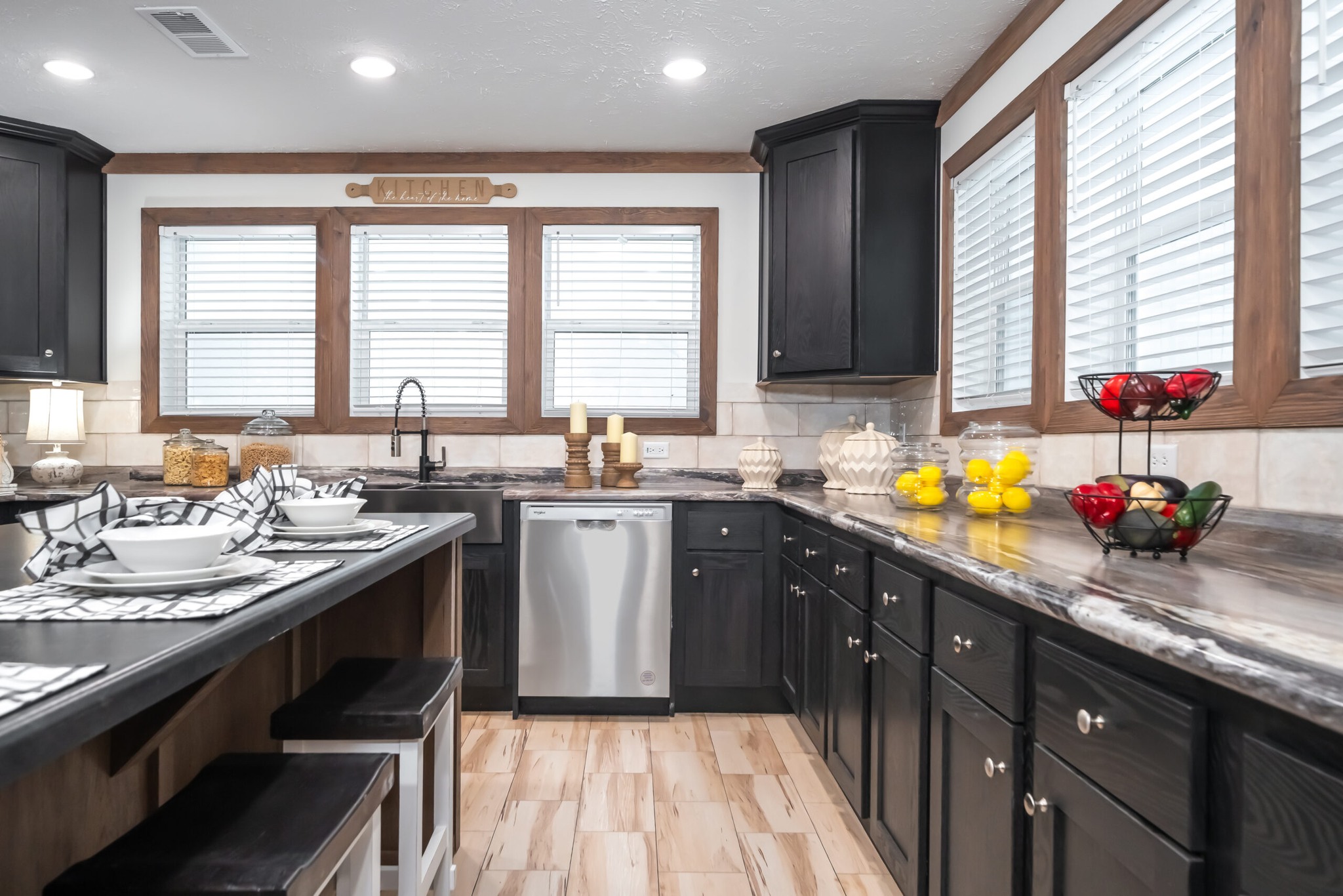 A modern kitchen with dark cabinets, light wood flooring, and a marble countertop. A fruit basket and candles add a cozy touch beneath large windows.