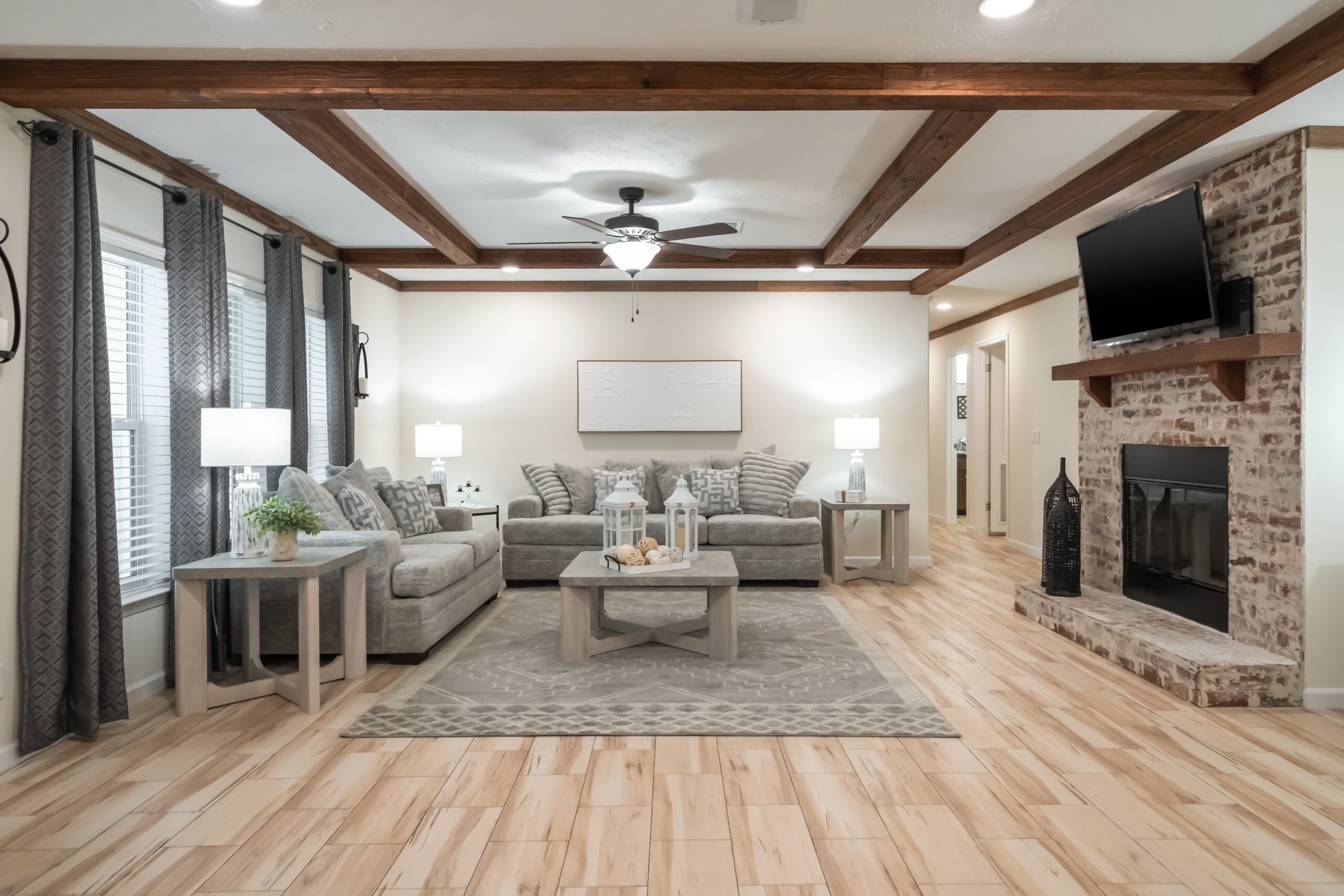 Spacious living room with wood beam ceiling, a gray sectional sofa, patterned pillows, light wood floor, and brick fireplace. Airy, calm ambiance.