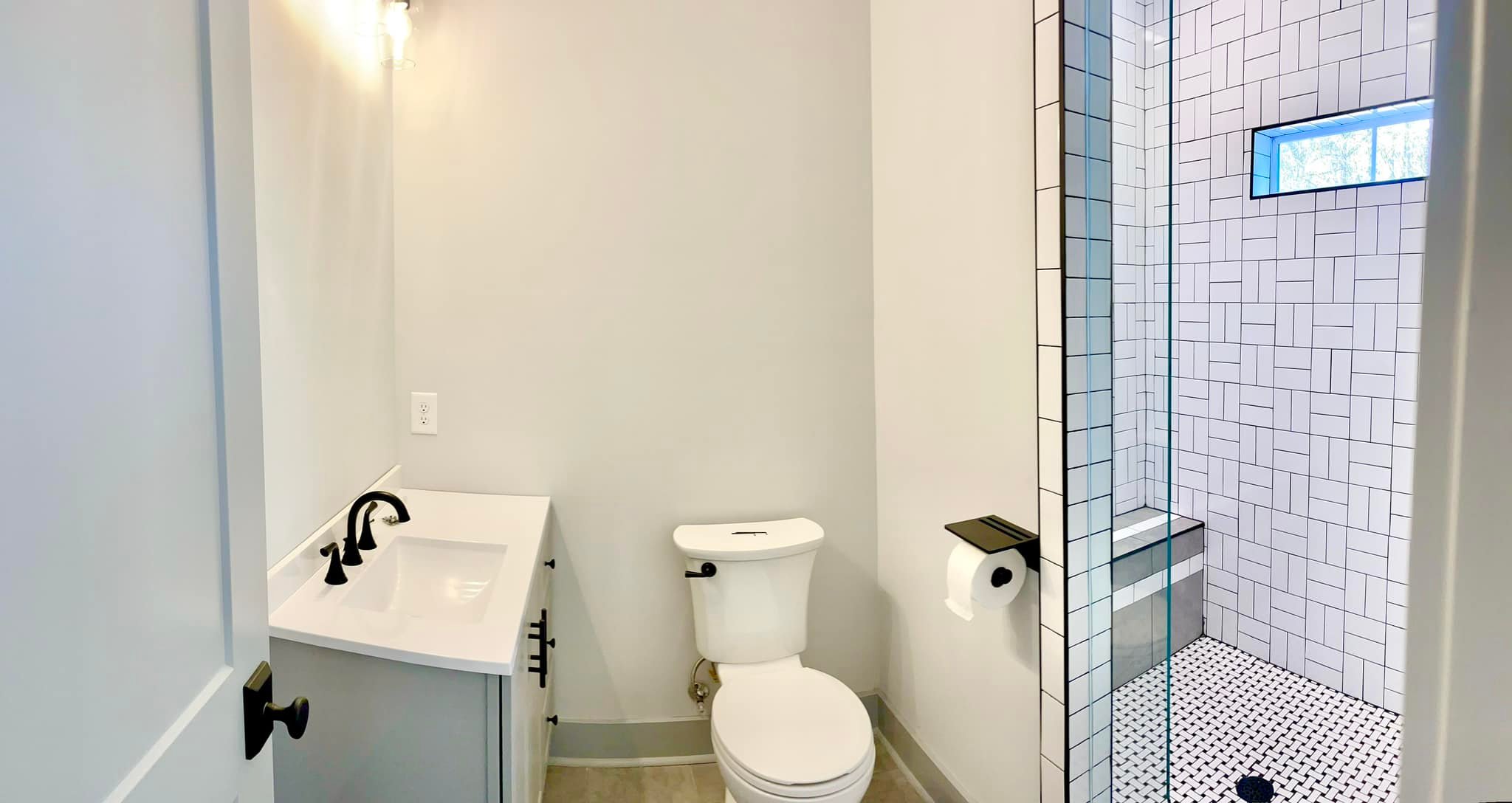 Small modern bathroom with white walls, featuring a white vanity with a black faucet, a toilet, and a shower with geometric tile and glass door. Bright and clean.