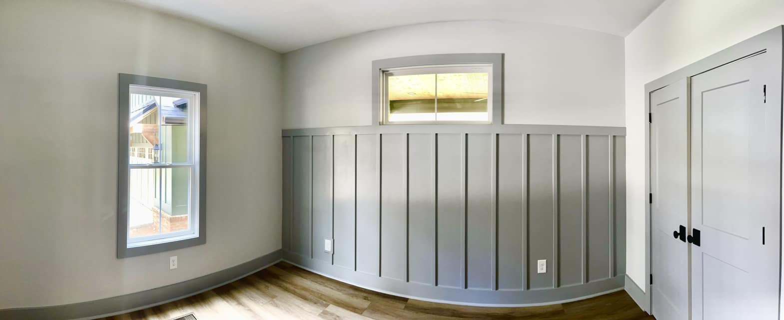 A bright, empty room with gray wainscoting, a light brown wood floor, and two windows—one larger and one smaller. The mood is clean and minimalistic.