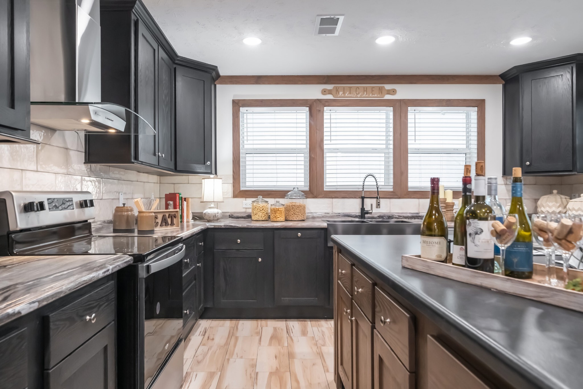 Modern kitchen with black cabinets and a marble countertop. Windows above the sink let in natural light. Bottles and corks on the island add a cozy touch.