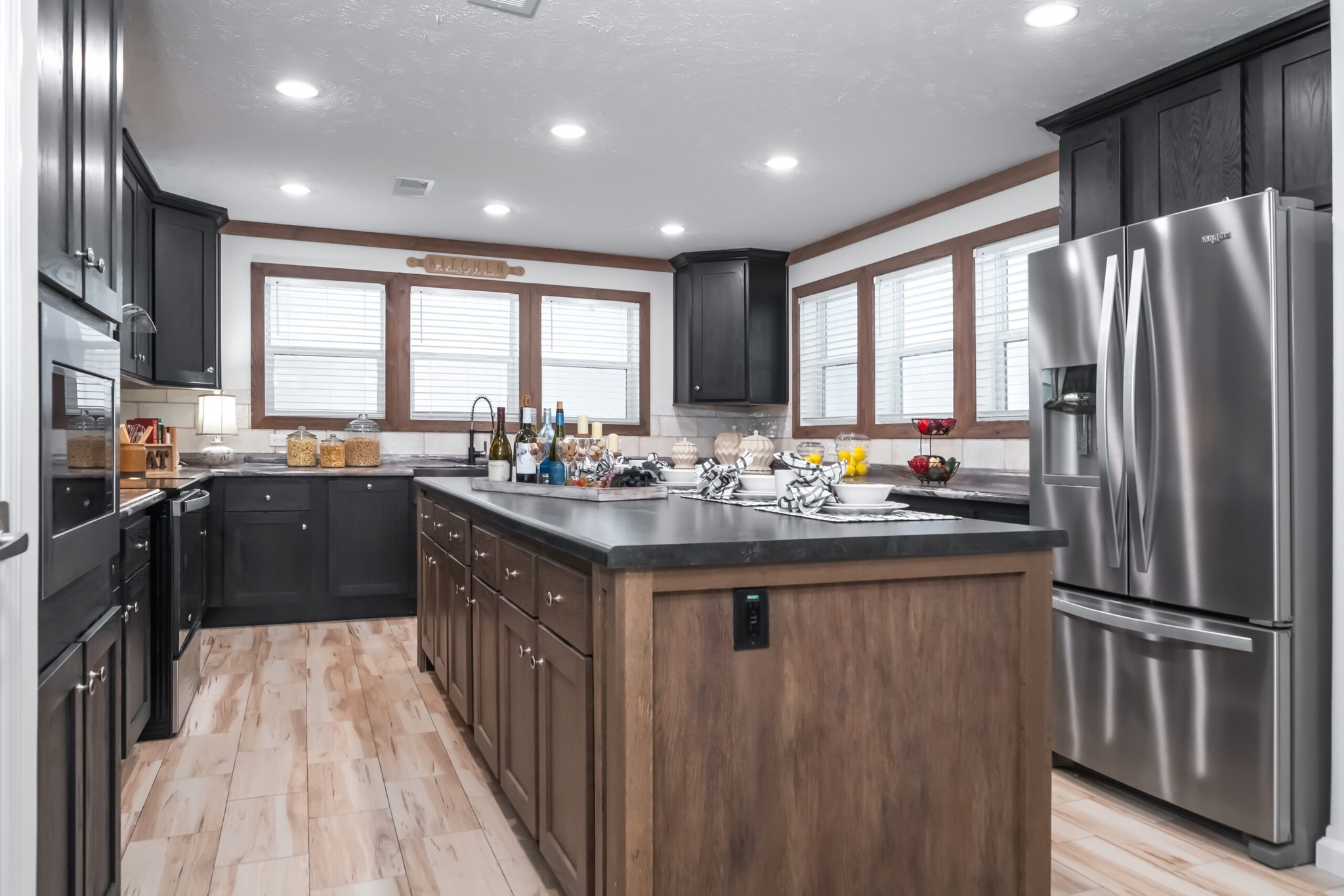 A modern kitchen with dark cabinets, stainless steel appliances, and a wooden island. Bright overhead lights and large windows create an inviting atmosphere.