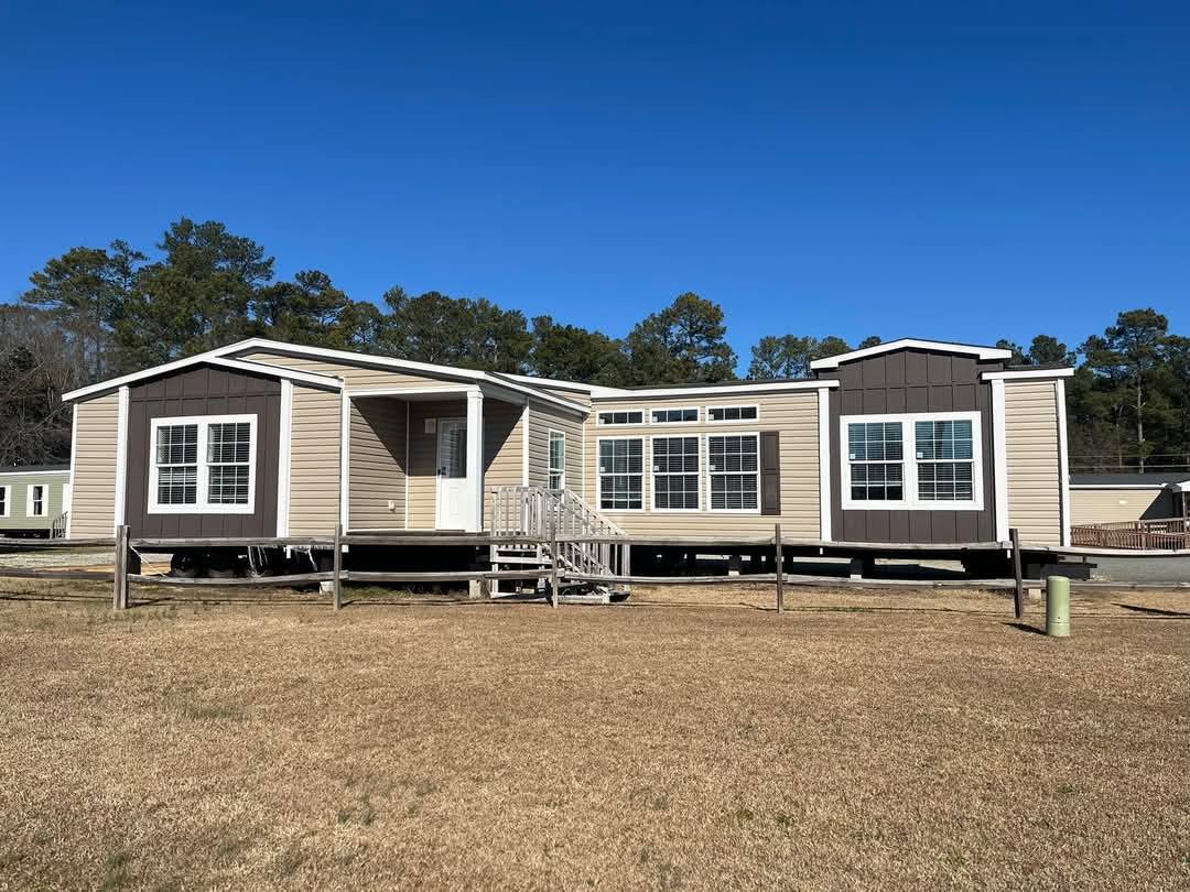A beige and brown modular home sits on a grassy lot under a clear blue sky. Large windows and a small porch are prominent, with trees in the background.