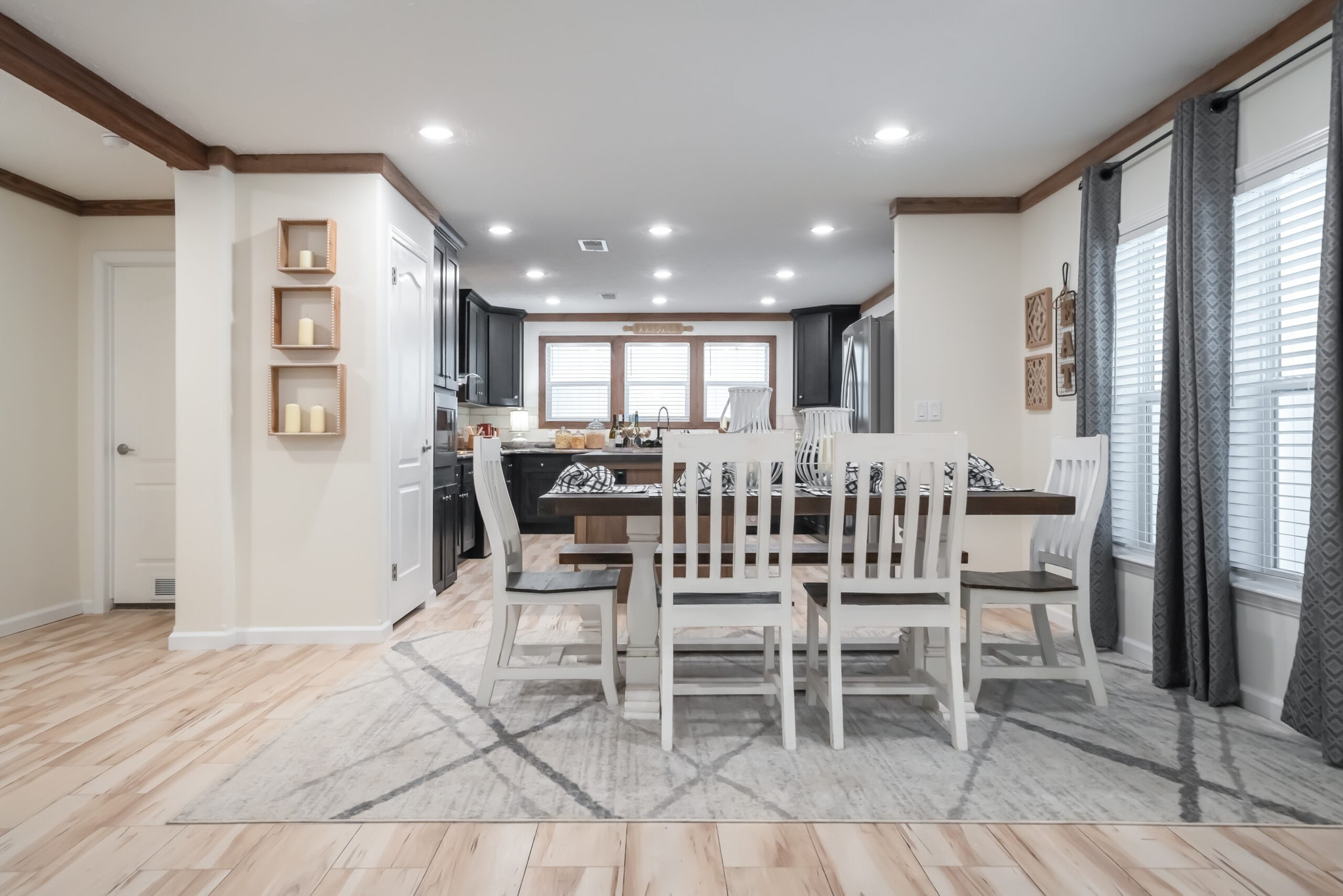 Bright dining room with a rustic wooden table, surrounded by six white chairs on a light patterned rug. Warm-toned floor, soft lighting, and large windows with gray curtains create an inviting atmosphere.