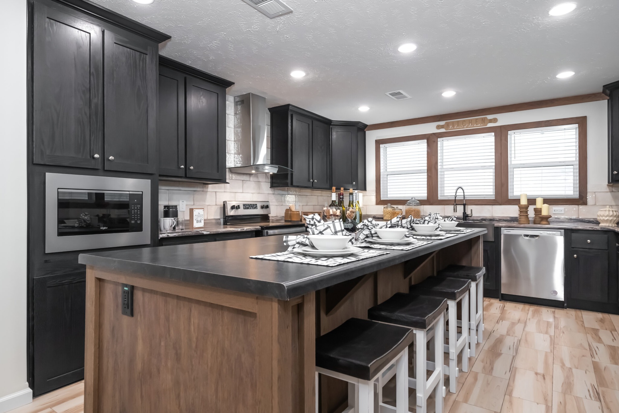 Modern kitchen with black cabinets, wooden island, and white stools. Bright lighting highlights neatly set tableware and stainless steel appliances. Cozy and inviting ambiance.