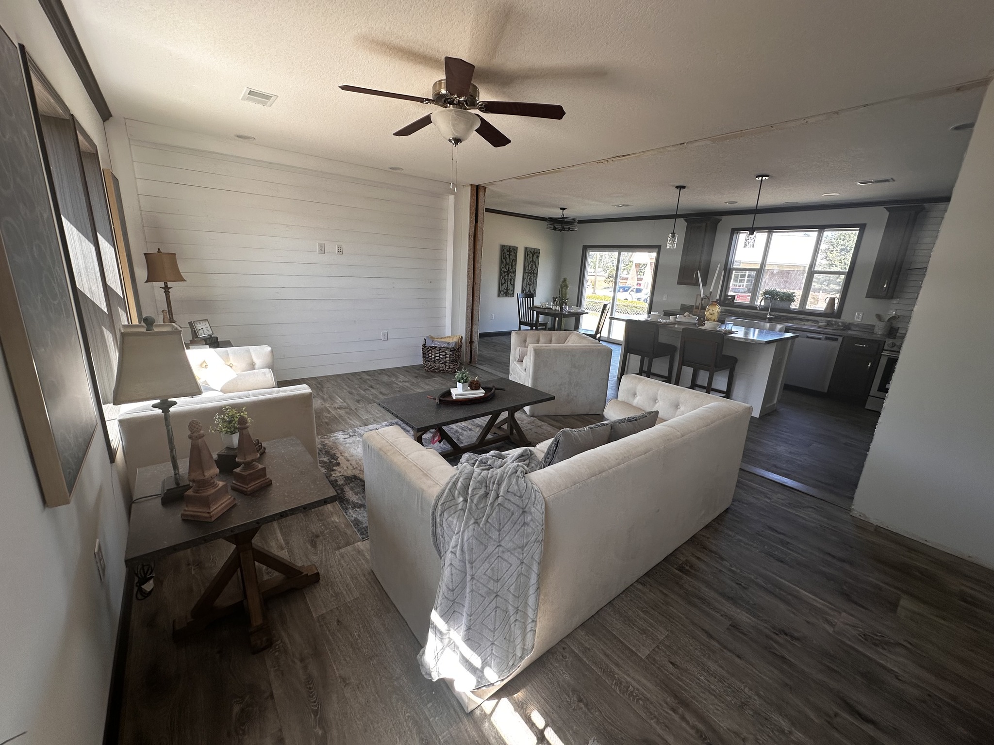 Spacious living area with beige couches, wooden tables, and a ceiling fan. Sunlit room with hardwood floors leads to a dining and kitchen area. Cozy and inviting atmosphere.
