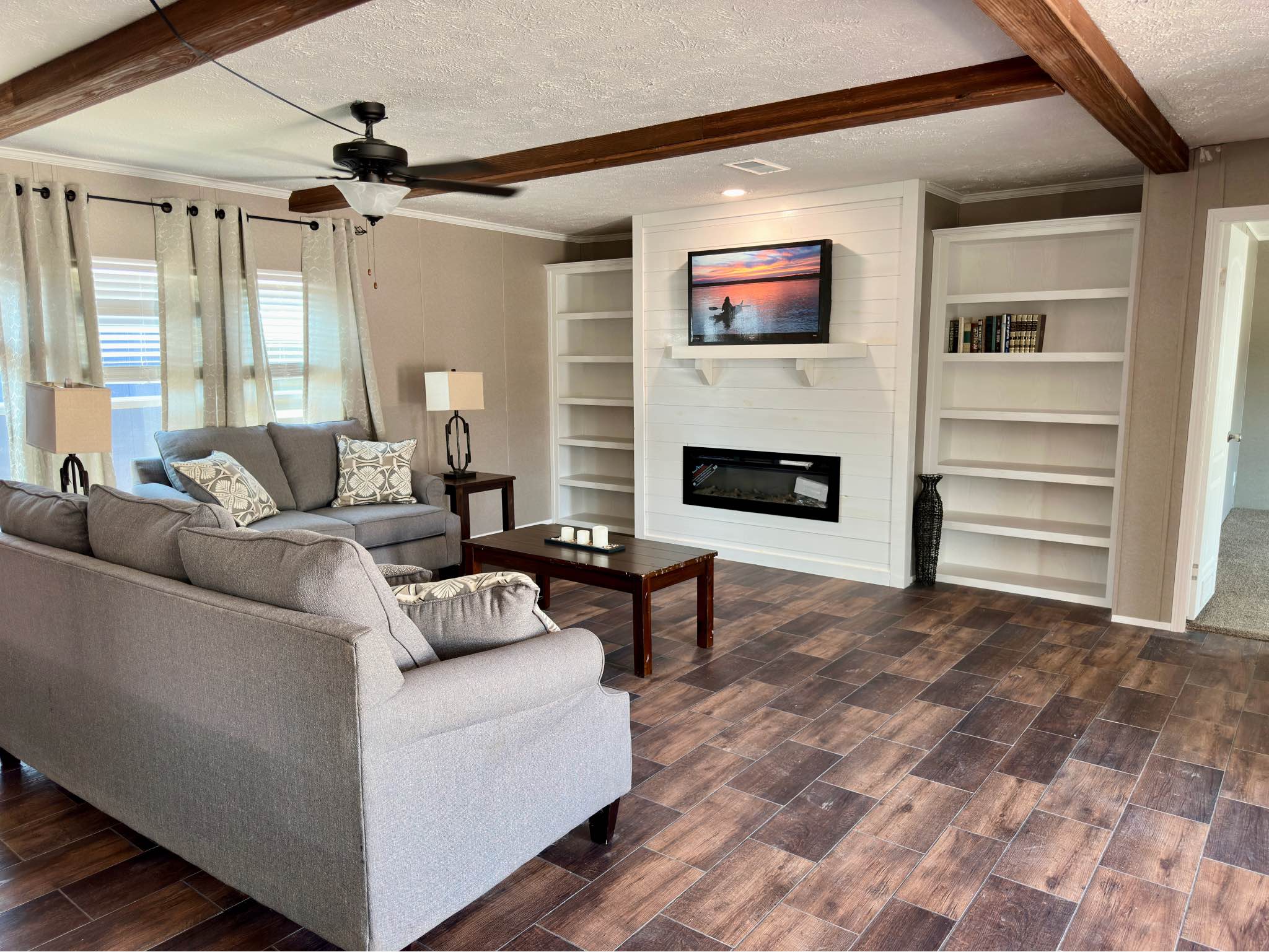 A cozy living room features a gray sectional sofa, wooden coffee table, and built-in shelves. A TV and fireplace are mounted on a shiplap wall.