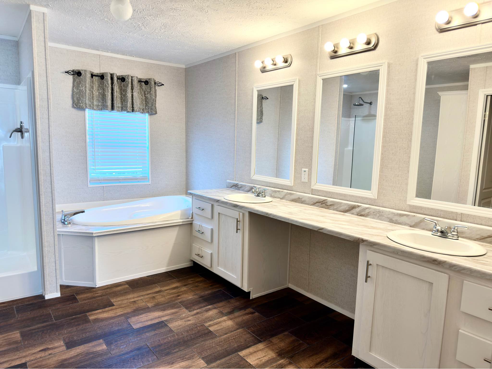 Spacious bathroom with dual sinks, marble countertop, and wooden floor. A white bathtub sits under a curtained window, lit warmly by overhead lights.