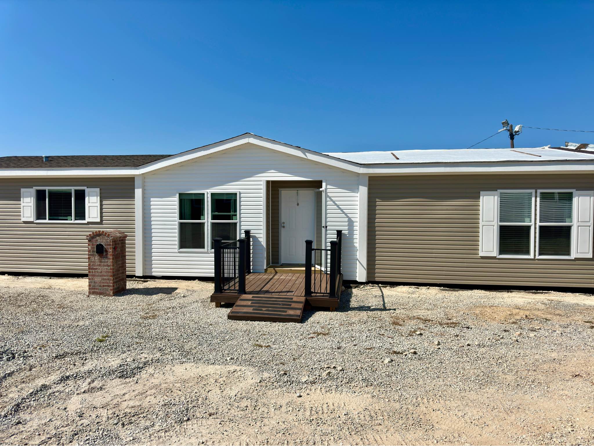 Single-story prefabricated home with a white and beige exterior under a clear blue sky. A small wooden ramp leads to the front door, set in a gravel yard.