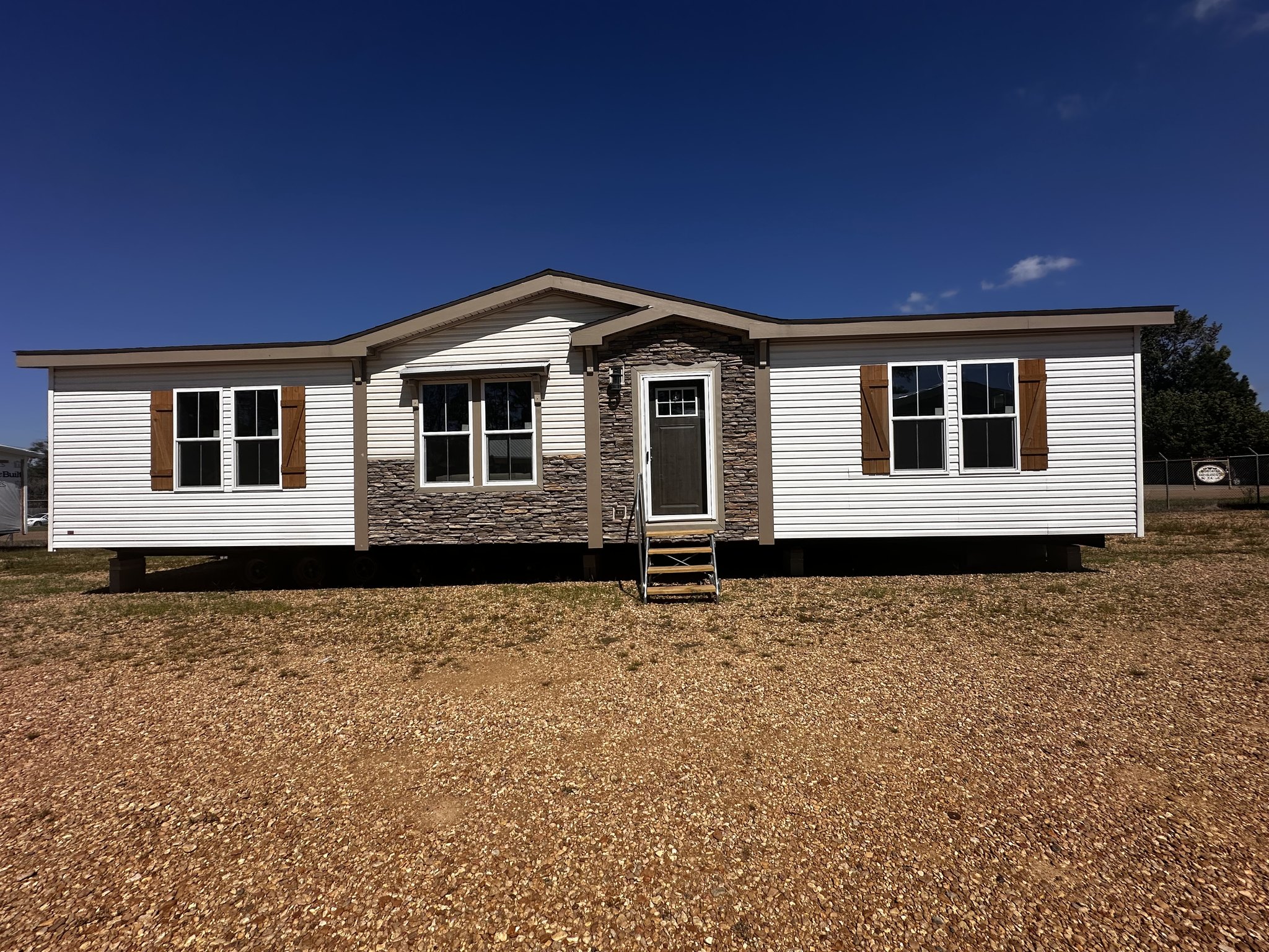 A modern manufactured home with white siding and stone accents stands on a gravel lot under a clear blue sky. Brown shutters frame the windows, creating a welcoming feel.