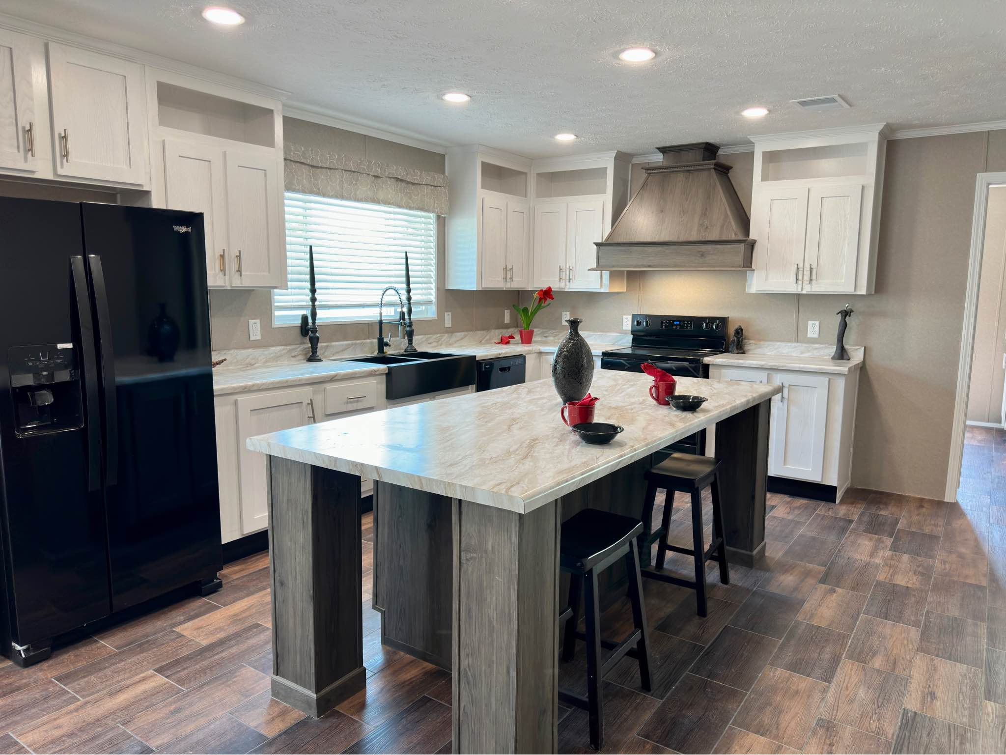 Modern kitchen with white cabinets, black appliances, and a light stone countertop island. Dark wood flooring and red utensils add warmth.