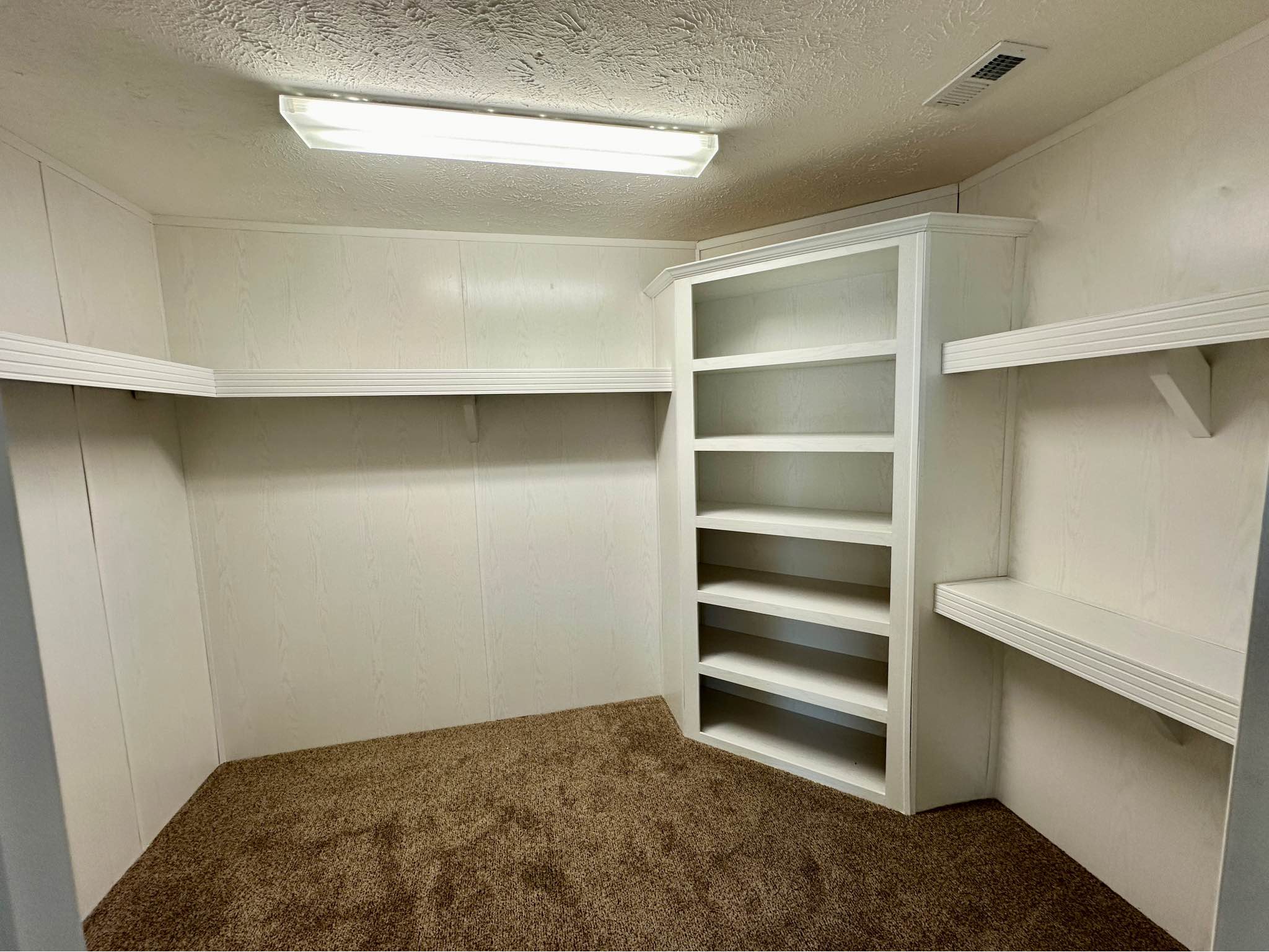 A small, carpeted walk-in closet with white walls and ceiling. The room features built-in shelves on two walls and a single fluorescent light overhead.