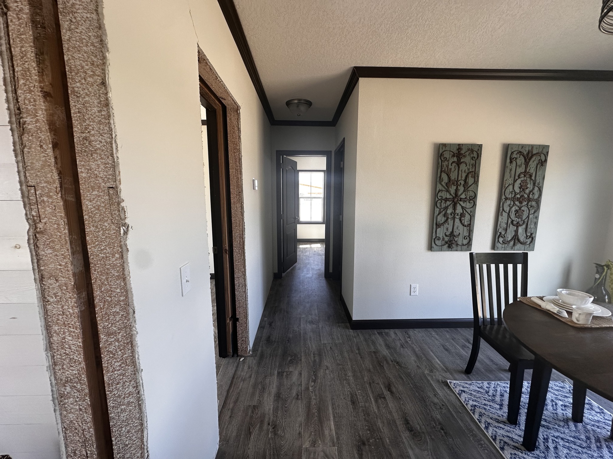 Hallway with wood flooring leading to a sunlit room. Dining table with place settings on the right, and decorative metal wall art enhancing a cozy atmosphere.