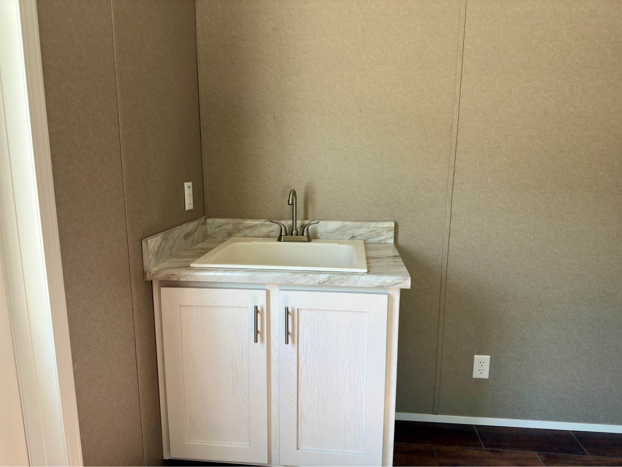 A small white sink with a silver faucet is set in a corner with a marble countertop and white cabinet. The walls are beige, and the flooring is dark brown.