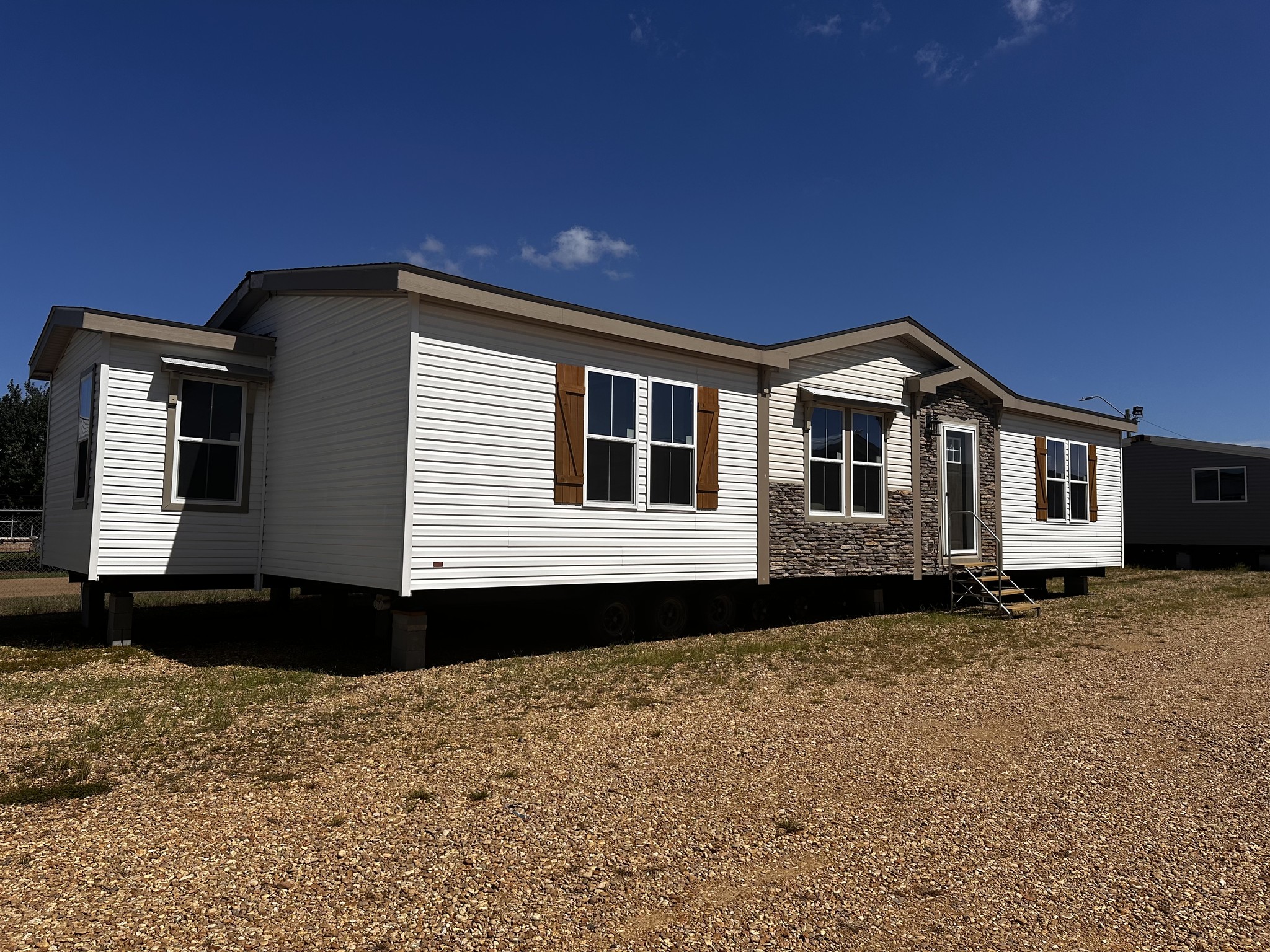 A white manufactured home with brown shutters and stone accents is set on a gravel lot under a clear blue sky, conveying a sense of simplicity and warmth.
