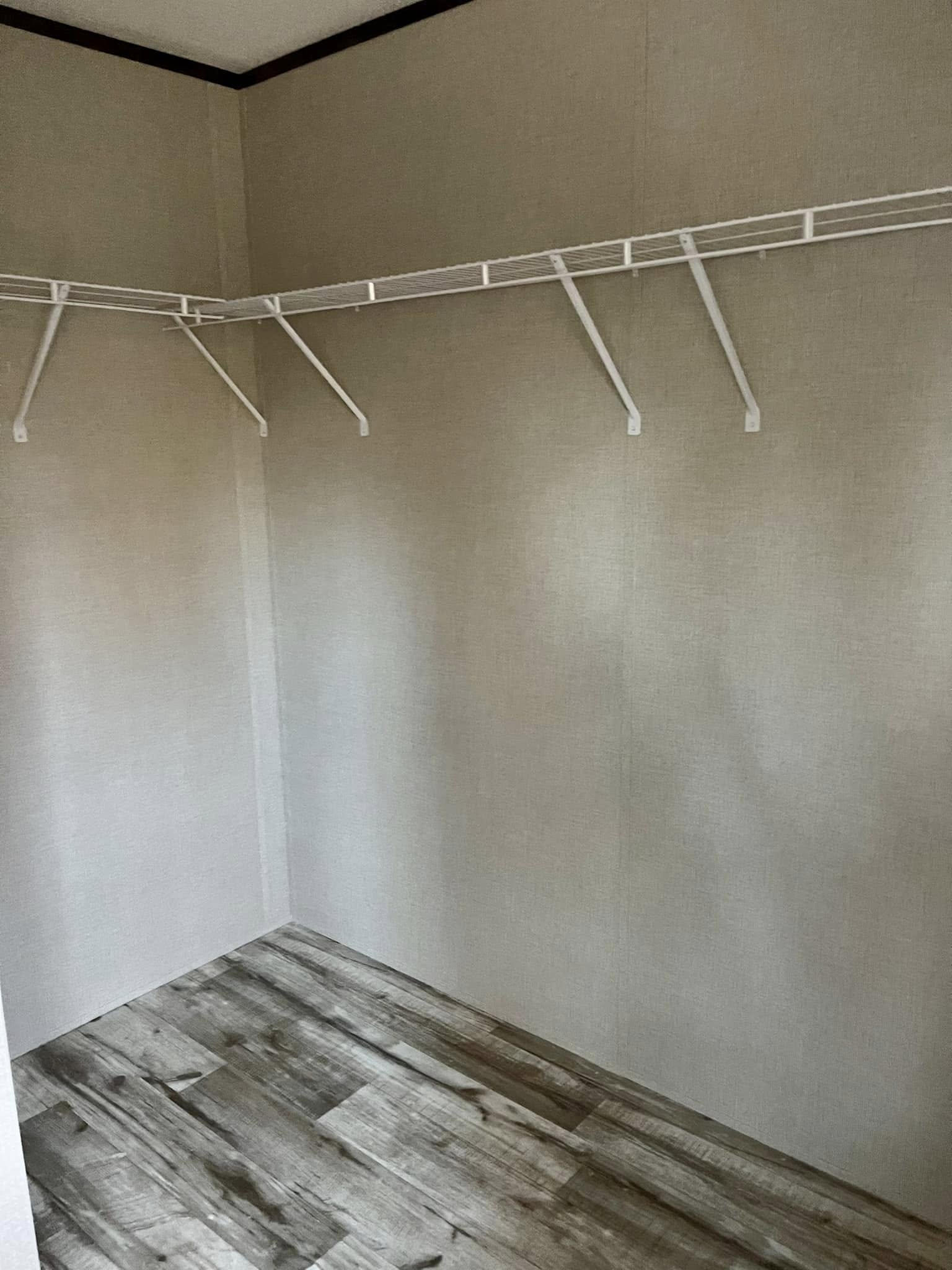 Empty walk-in closet with beige walls and rustic wood flooring. A white wire shelf runs along the walls, creating a minimalist, clean atmosphere.