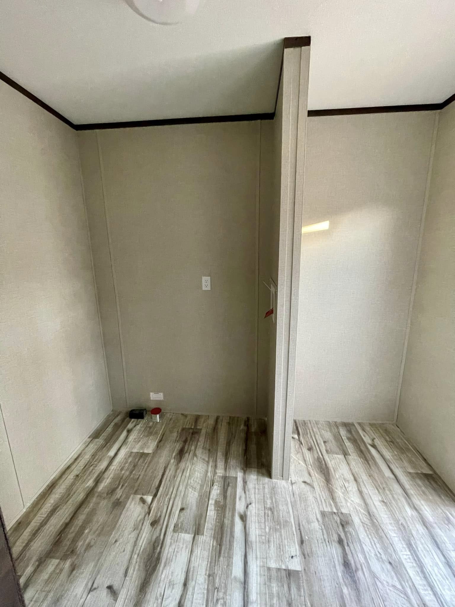 Empty room with light wood laminate flooring, beige walls, and a partition dividing the space. A small light switch is mounted on the far wall.