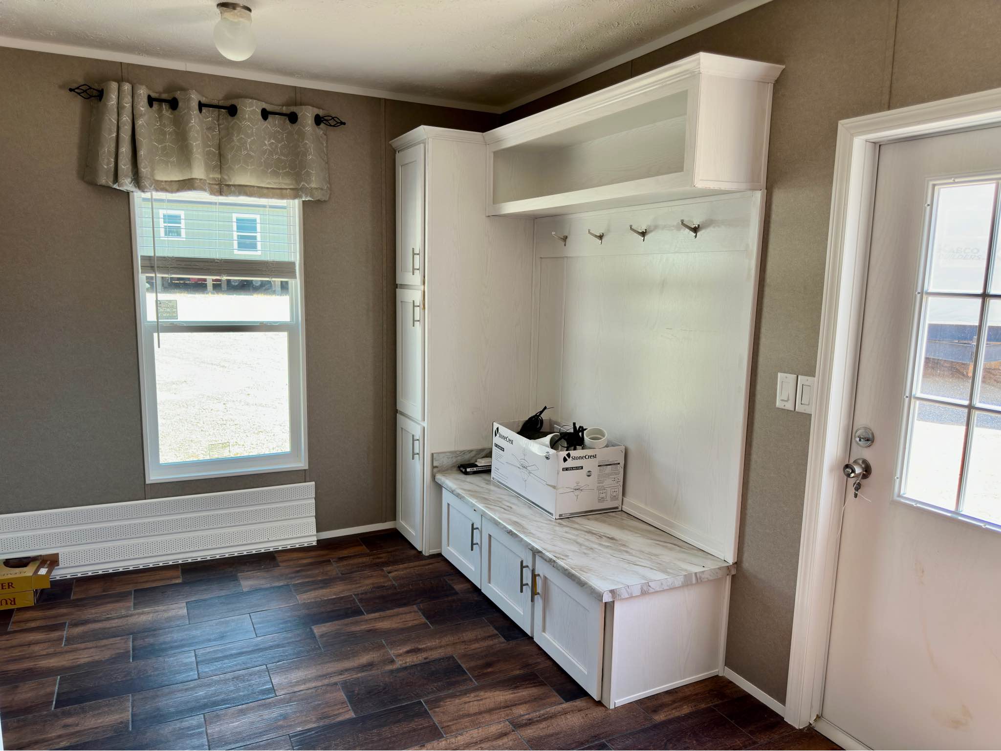 Bright entryway with wood flooring, featuring white cabinetry with hooks and a bench. A window with curtains lets in light. A cardboard box sits on the bench.