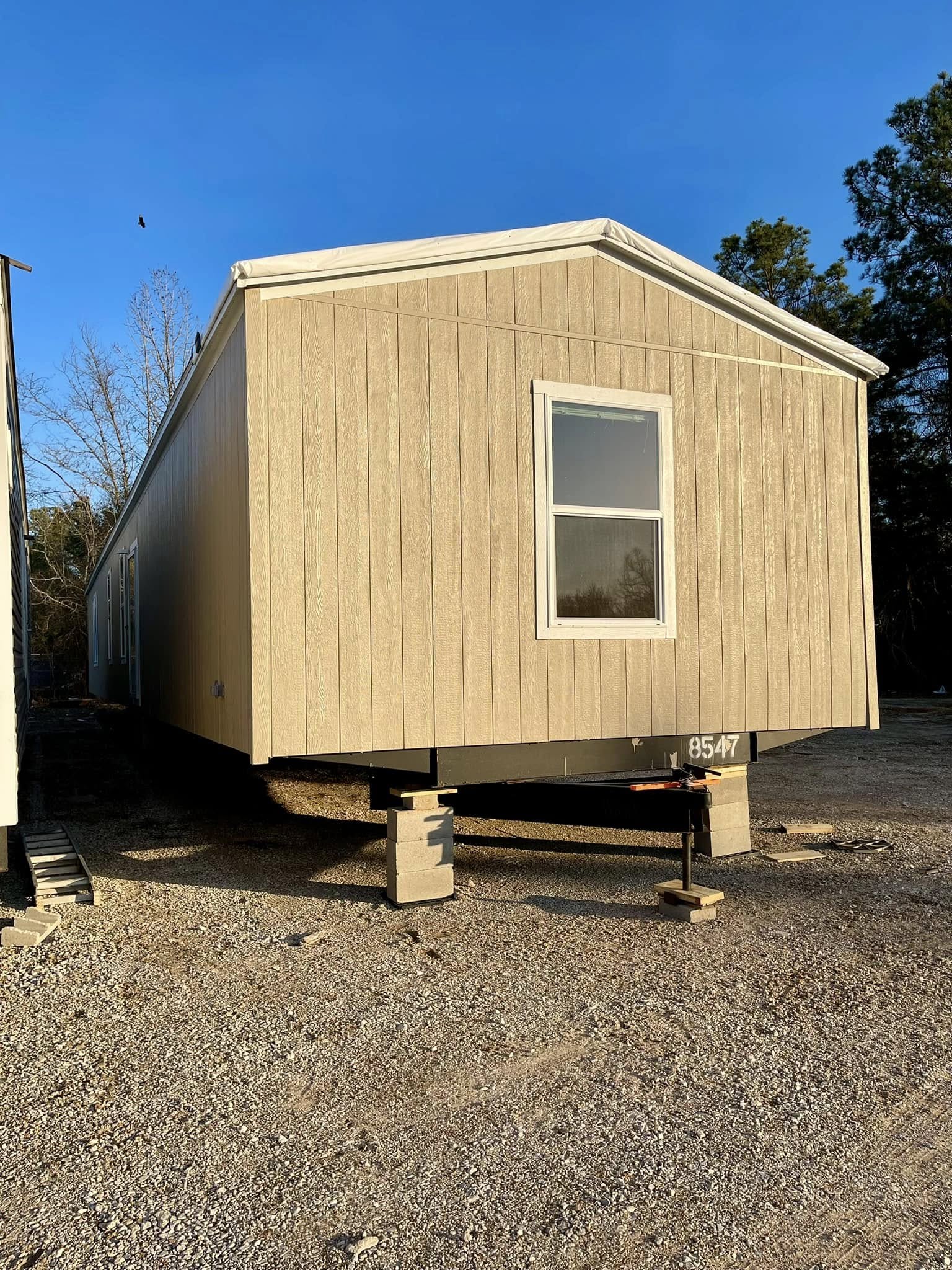 Beige mobile home on concrete blocks in a gravel lot, bathed in warm sunlight. Surrounded by trees under a clear blue sky, conveying a peaceful tone.