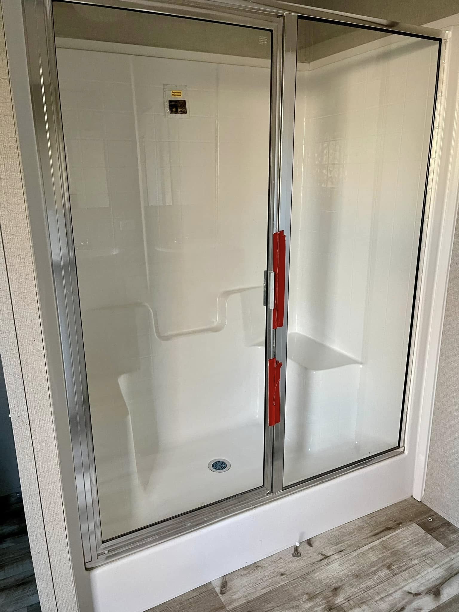 A clean, modern shower with glass sliding doors outlined in a metal frame. Red tape is attached to the handles. The interior is white with a small bench.