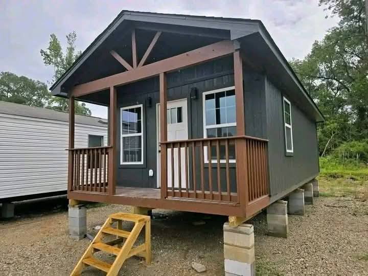 Small dark gray cabin with a brown wooden porch, elevated on concrete blocks. Surrounded by trees under a cloudy sky, conveying a rustic, serene vibe.