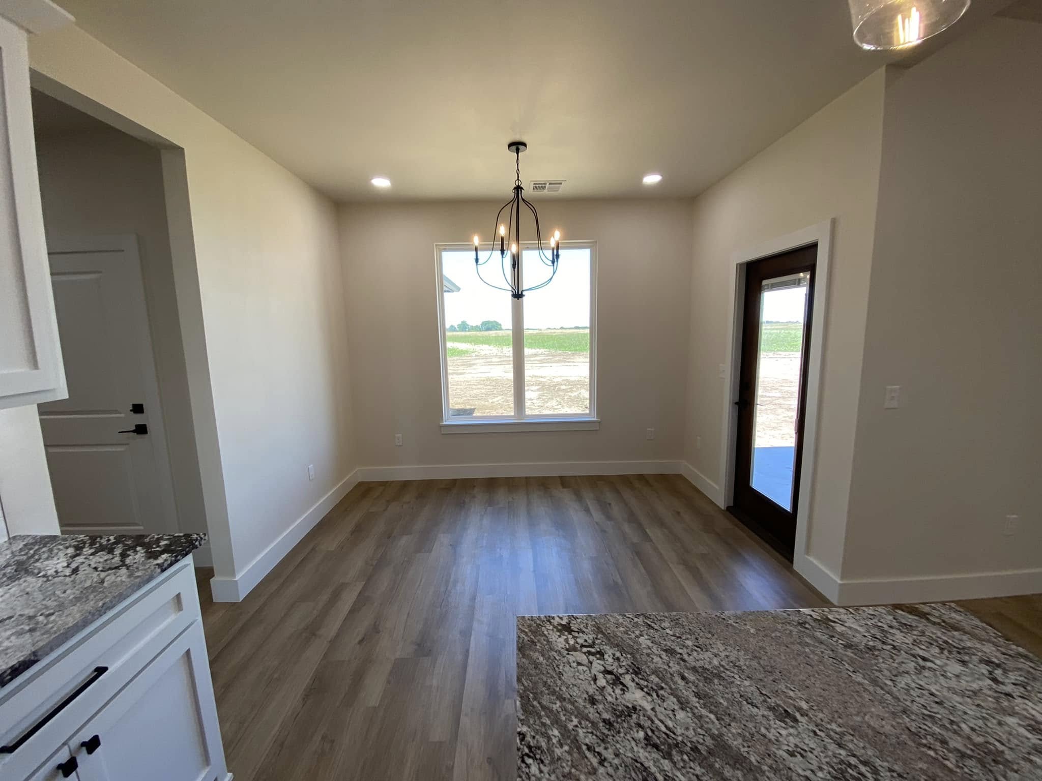 Bright dining area with wood flooring, a large window showing fields, recessed lighting, dark framed doors, granite counters, and a modern chandelier.