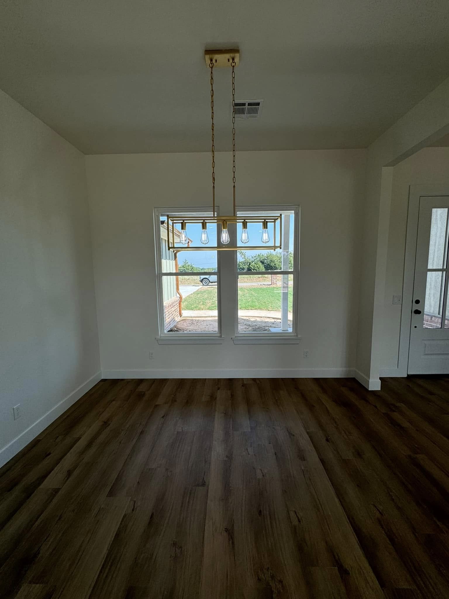 Empty room with light wood flooring, white walls, and a large window overlooking a grassy yard. A modern gold light fixture hangs from the ceiling.