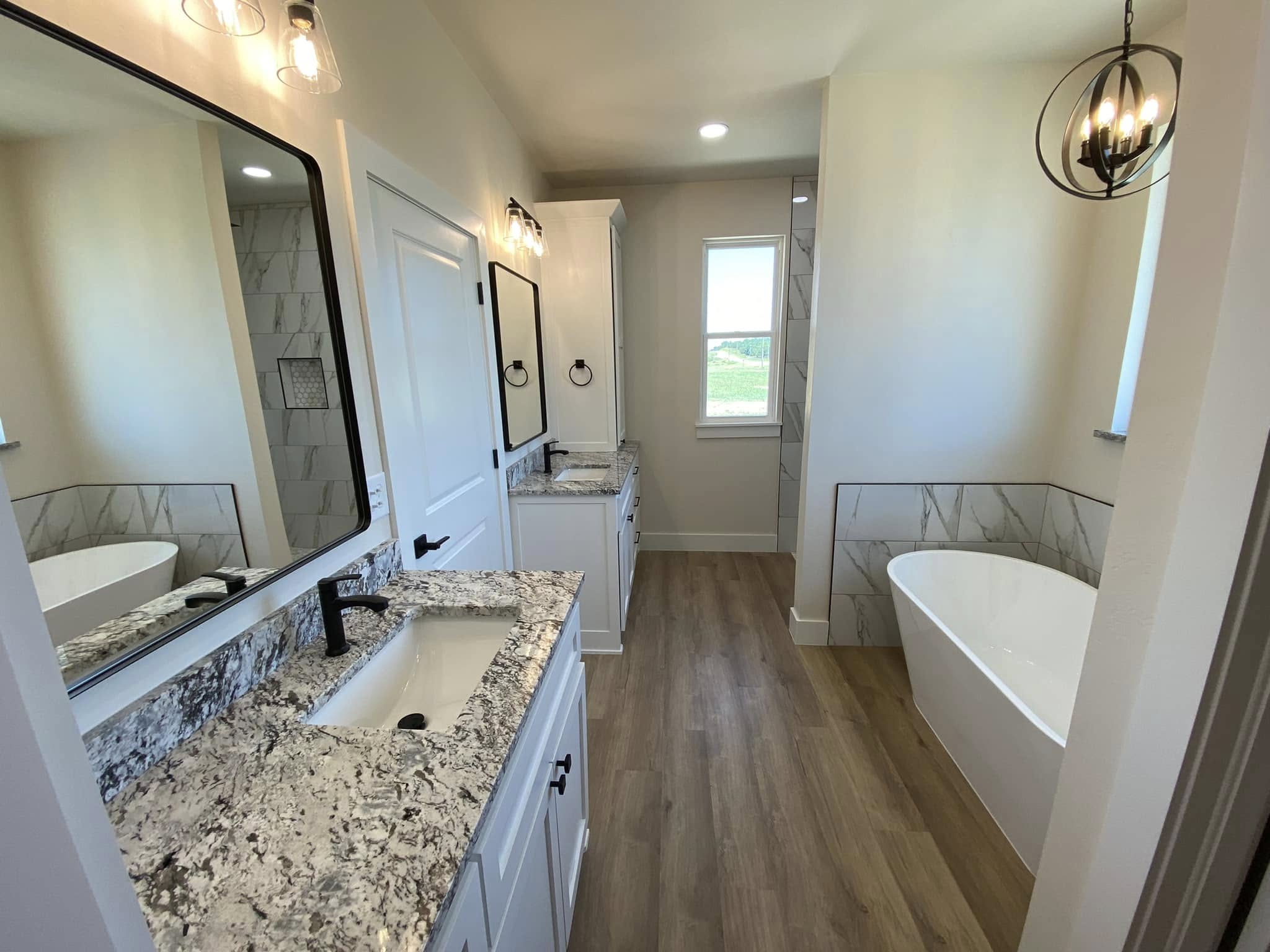 Modern bathroom with dual granite-topped vanities, black fixtures, and large mirrors. Freestanding tub near the window, wood flooring, neutral tones.
