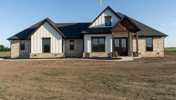 A modern, single-story house with a mix of white siding and beige brick exterior, large windows, and a dark roof. It's surrounded by a barren plot of land under a clear blue sky.
