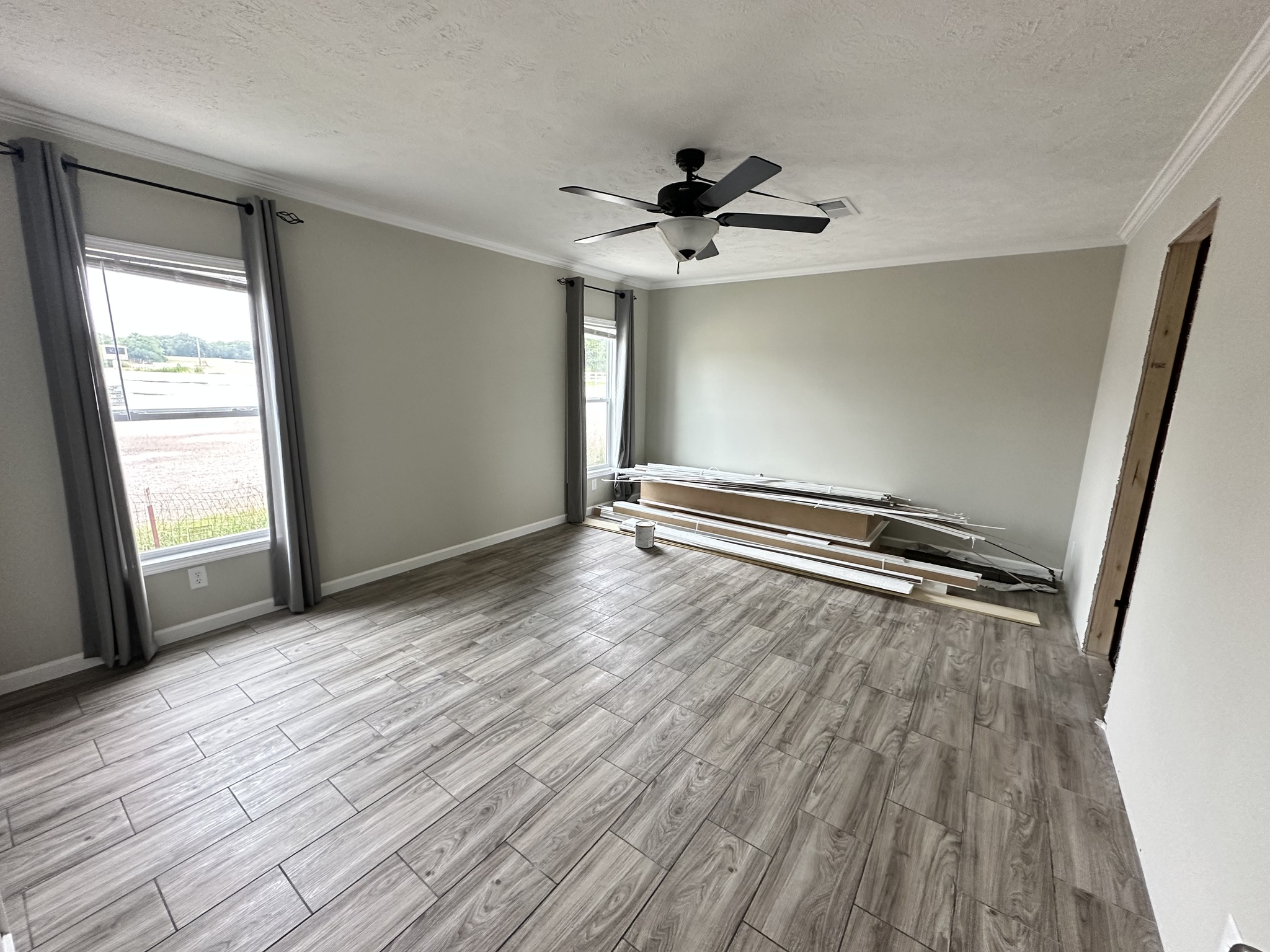 Empty room with light gray walls and wood-look tile floor. Two windows with gray curtains, a ceiling fan, and construction materials stacked by the wall.