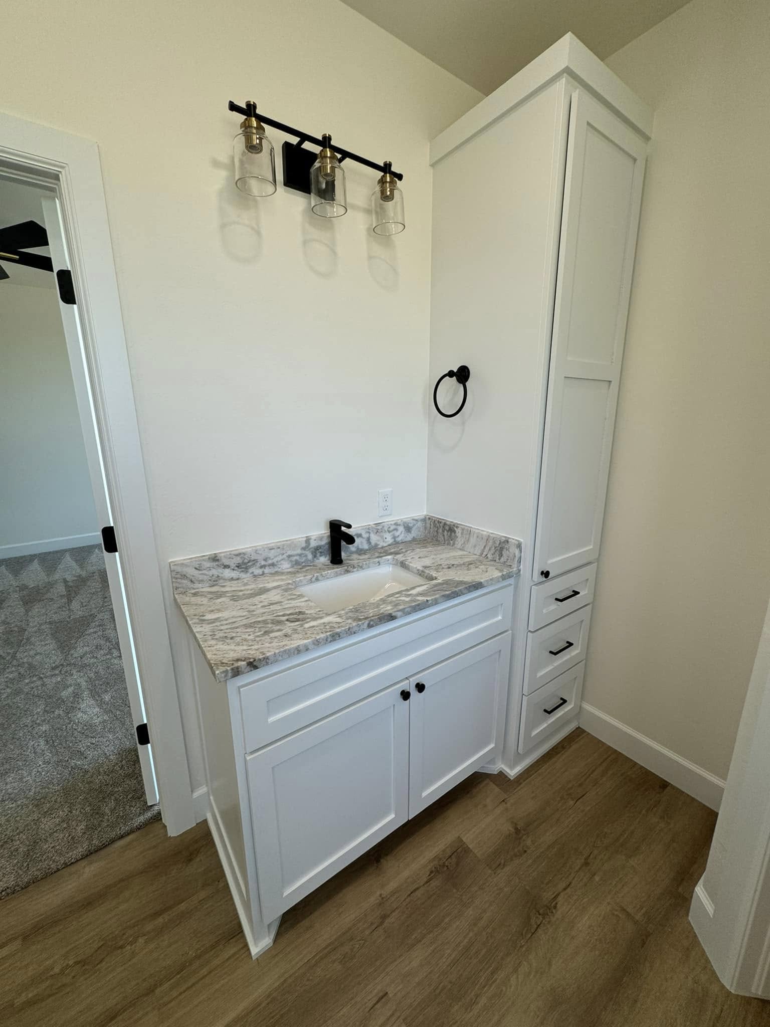 White bathroom vanity with a marbled countertop, black faucet, and vertical cabinet. Above are three bulb fixtures. Wood floor and plain walls add a modern touch.