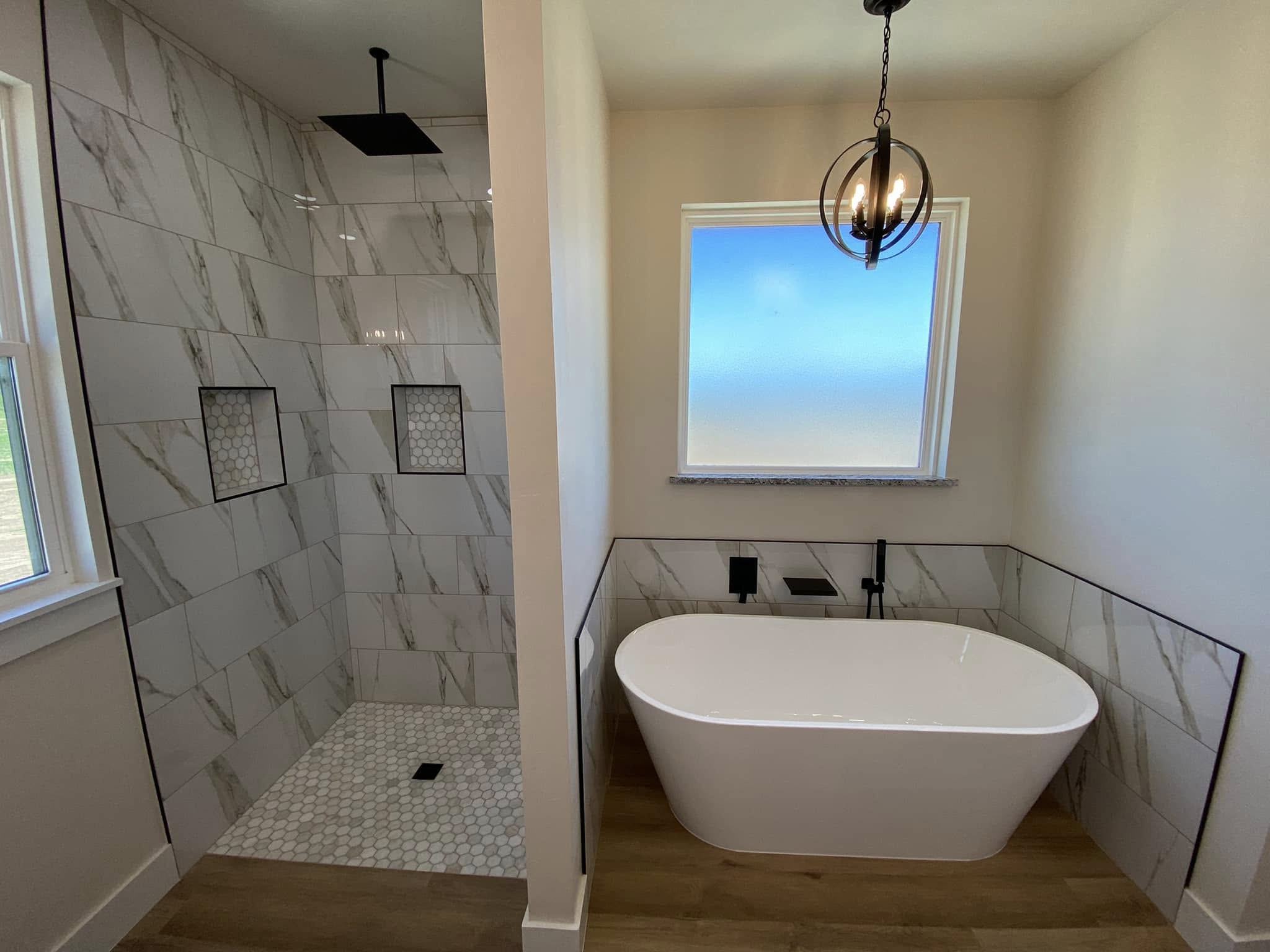 Modern bathroom with a marble-tiled shower and a freestanding white tub. A black pendant light hangs above, with a window casting soft light.