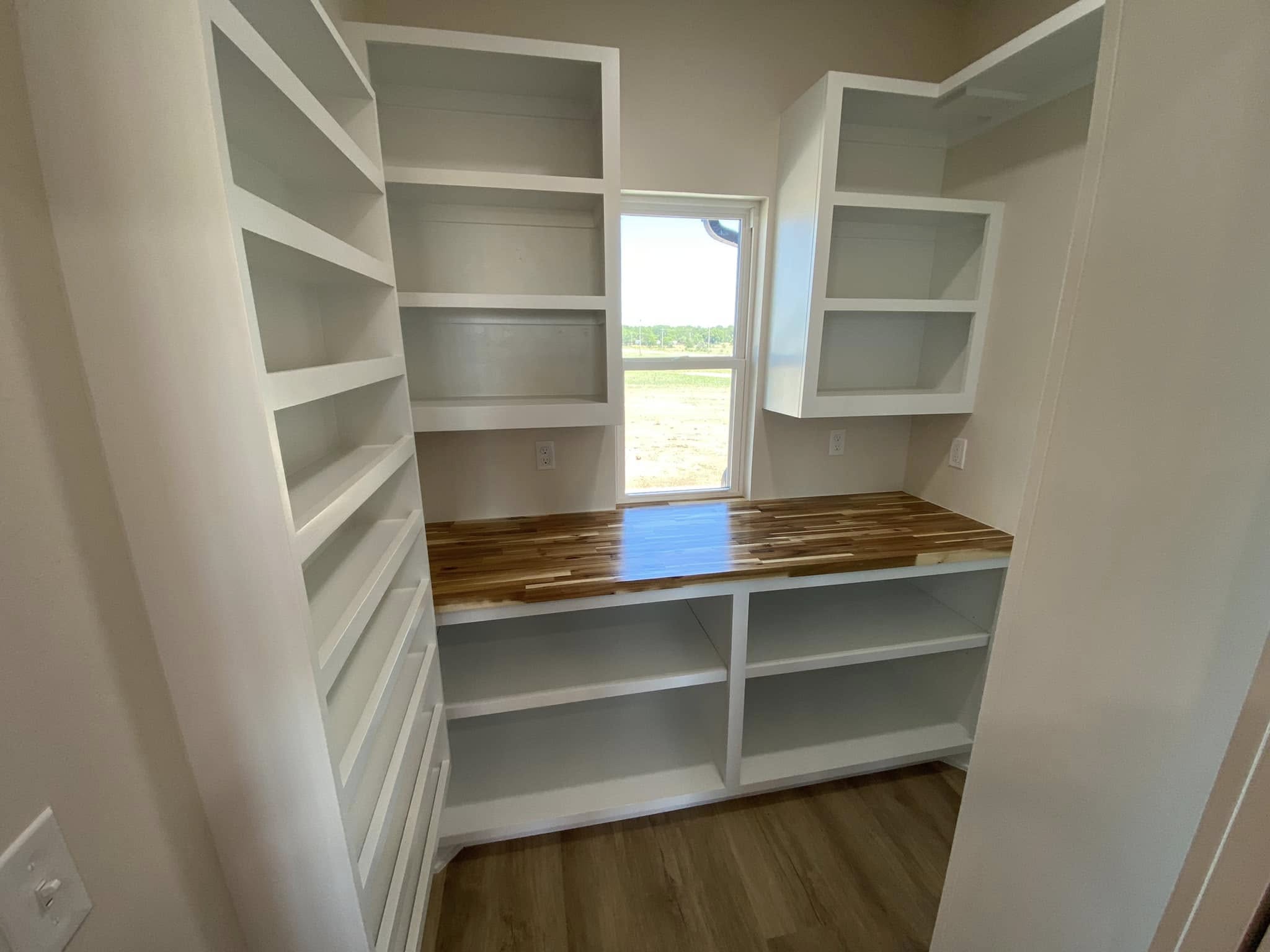 Small pantry with white shelving on both sides and a wood countertop beneath a window. The room has a clean, bright, and organized feel.