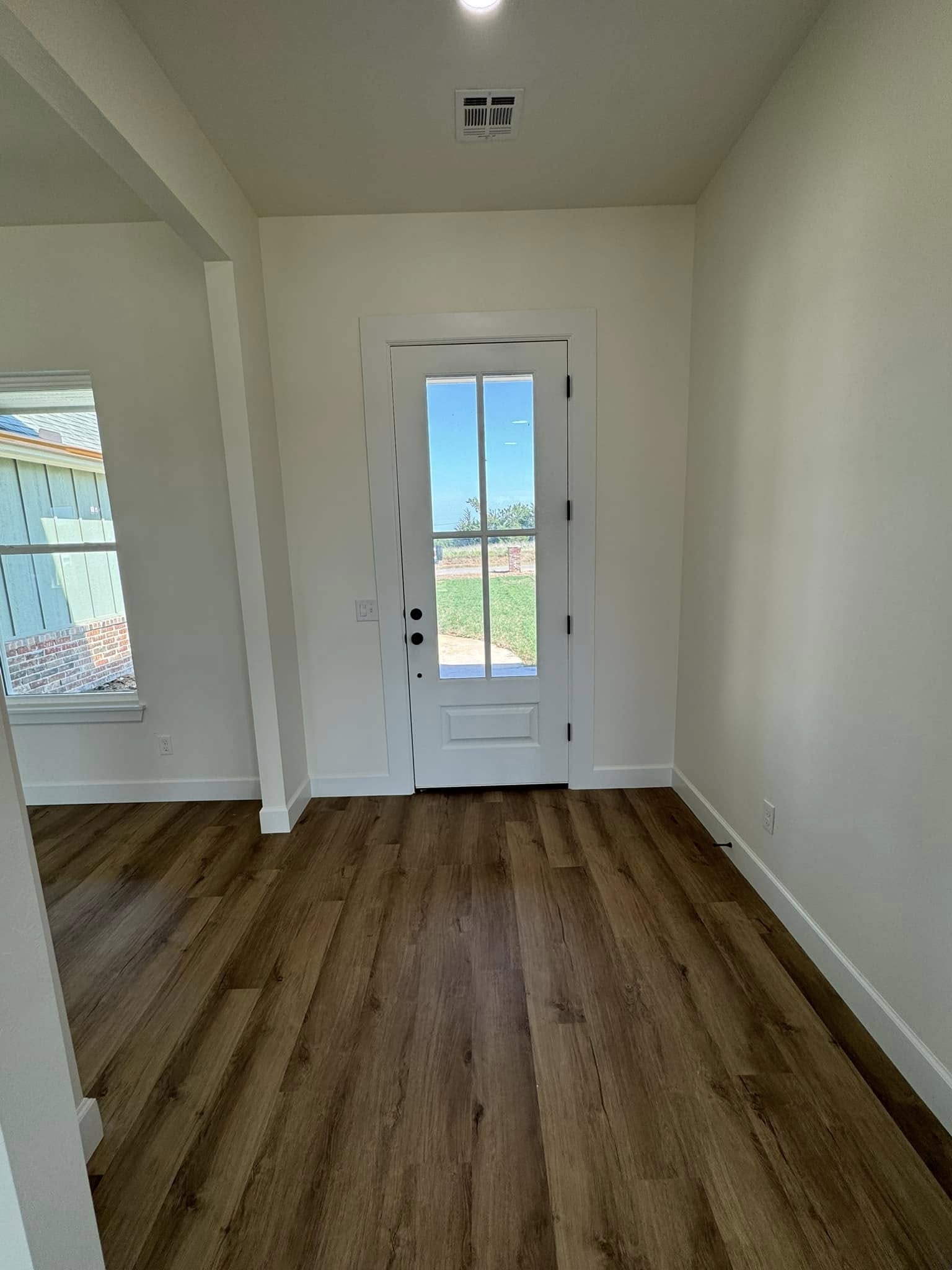 A bright entryway with a wooden floor leads to a white door with glass panes. Sunlight streams in, revealing a glimpse of grass outside. Calm and inviting atmosphere.