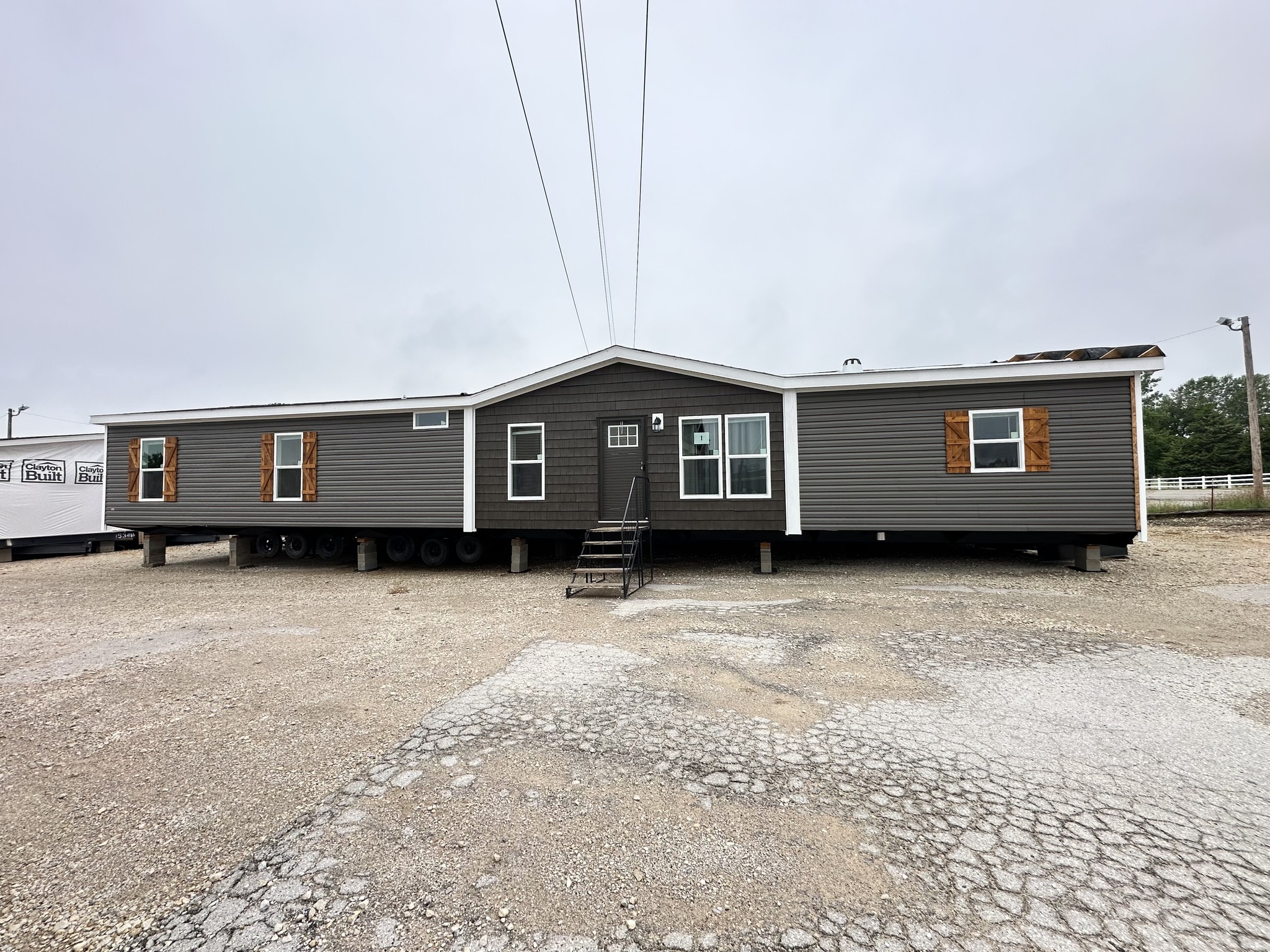 A grey manufactured home with white trim and wooden window shutters sits elevated on a gravel lot. Stairs lead to the front door. It's overcast.