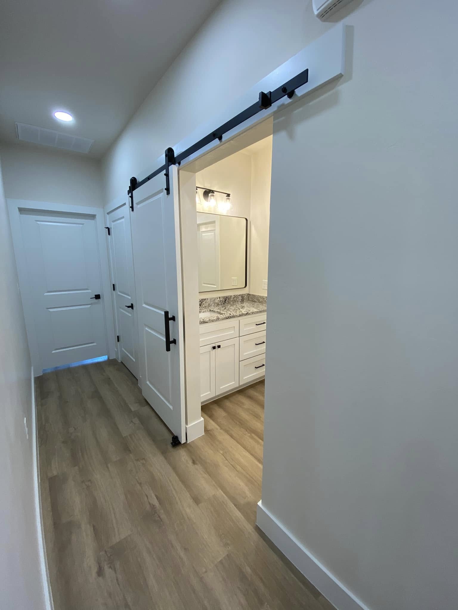 A modern hallway with light wooden flooring leads to a bathroom. A sliding barn door is open, showing a vanity with a large mirror and granite countertop, under soft lighting.