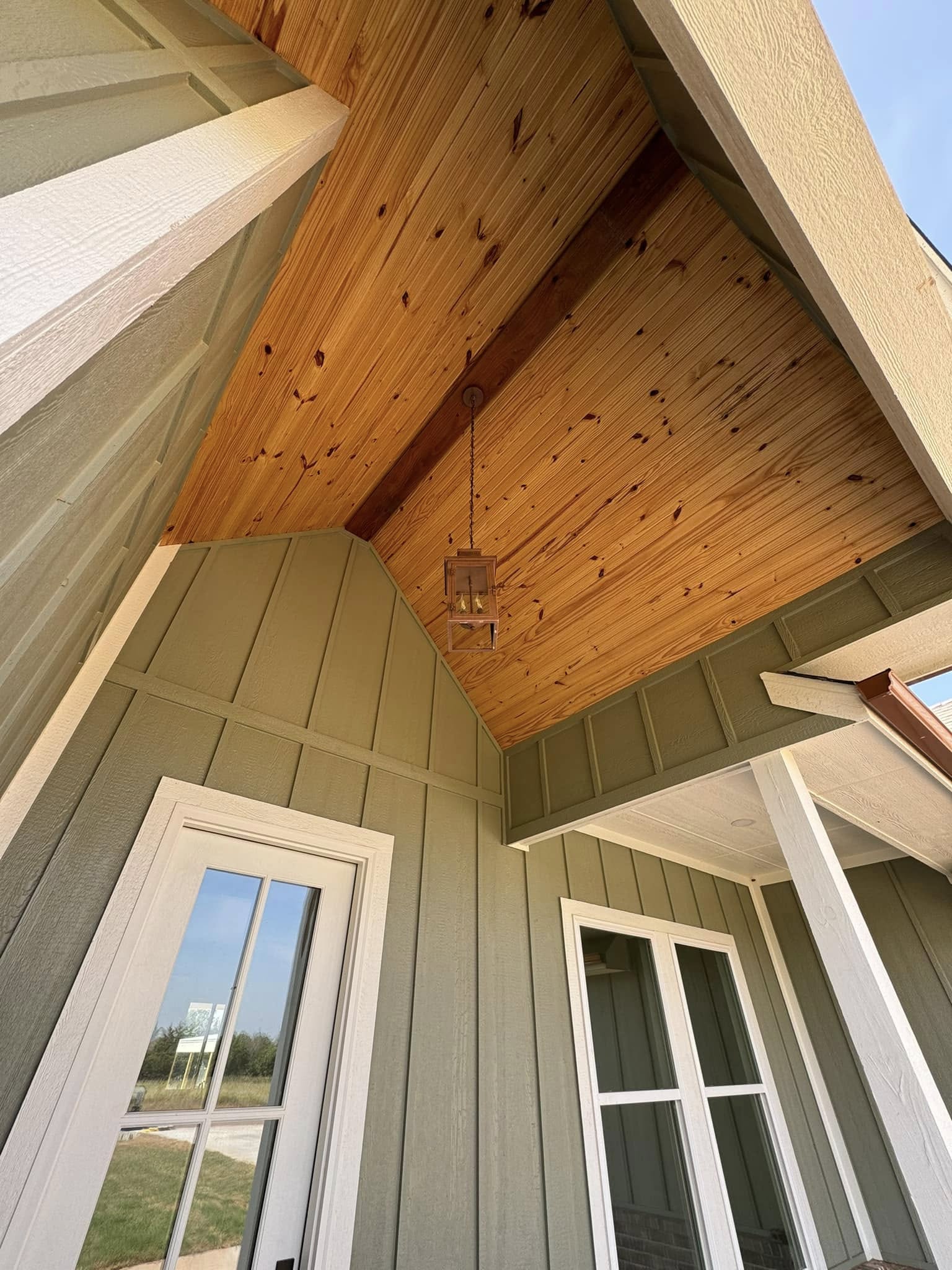 Rustic porch with wooden ceiling and green-painted siding, featuring large windows and a hanging lantern. Bright, sunny day creates a welcoming vibe.