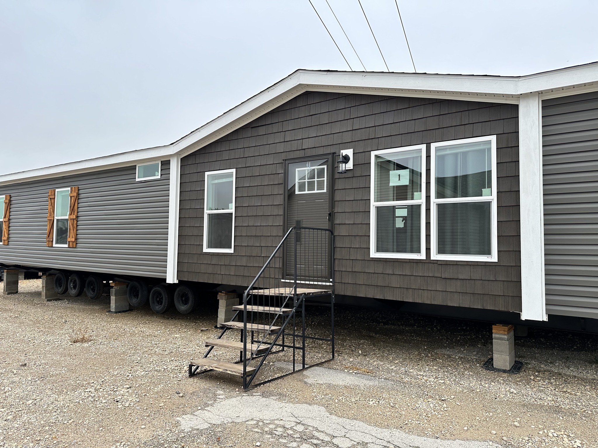 A large manufactured home on a gravel lot, featuring brown siding, white trim, and two windows. Metal steps lead to the front door. Overcast sky.