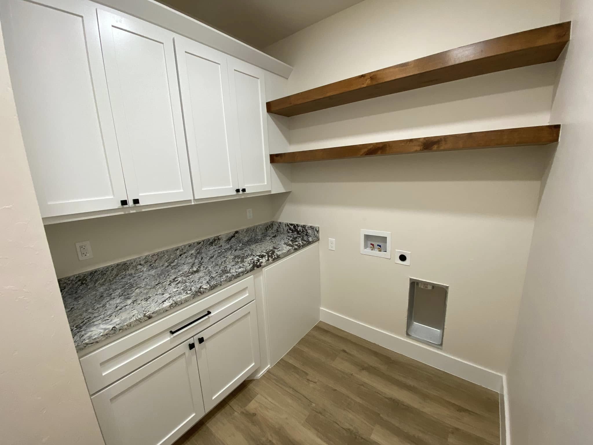 Small laundry room with white cabinets, granite countertop, and two wooden shelves on the wall. Empty appliance spaces on the right wall.