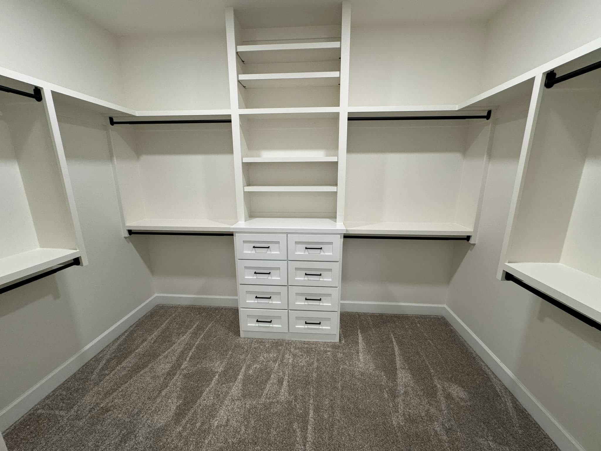 Minimalist walk-in closet with white shelves, multiple hanging rods, and a set of drawers on beige carpet, creating an organized and spacious feel.
