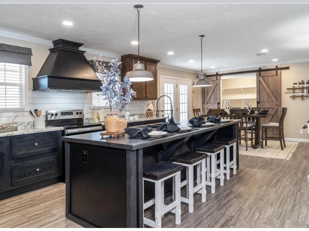 Modern kitchen with dark cabinets, marble countertops, and a central island with stools. Pendant lights hang overhead. Cozy dining area with sliding barn doors.