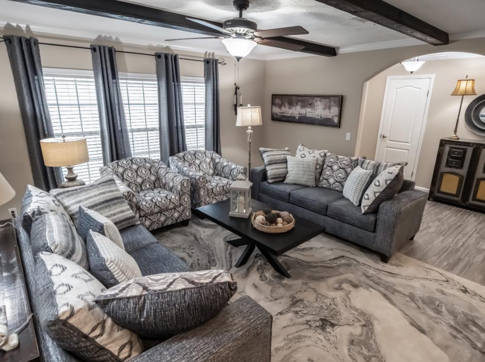A cozy living room with gray sofas and patterned armchairs arranged around a black coffee table. Large windows with gray curtains let in natural light. A ceiling fan with lights hangs above. Neutral tones create a warm, inviting atmosphere.