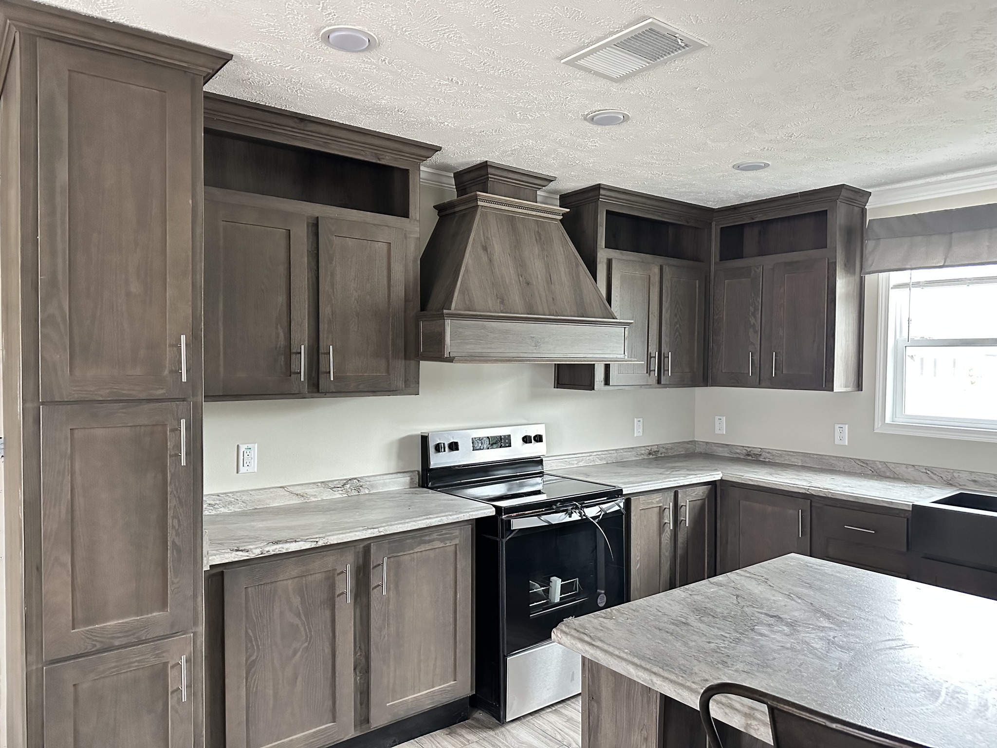Spacious kitchen with brown wooden cabinets, marble countertops, and a center island. Features a stainless steel stove and a large range hood. Bright, modern ambiance.