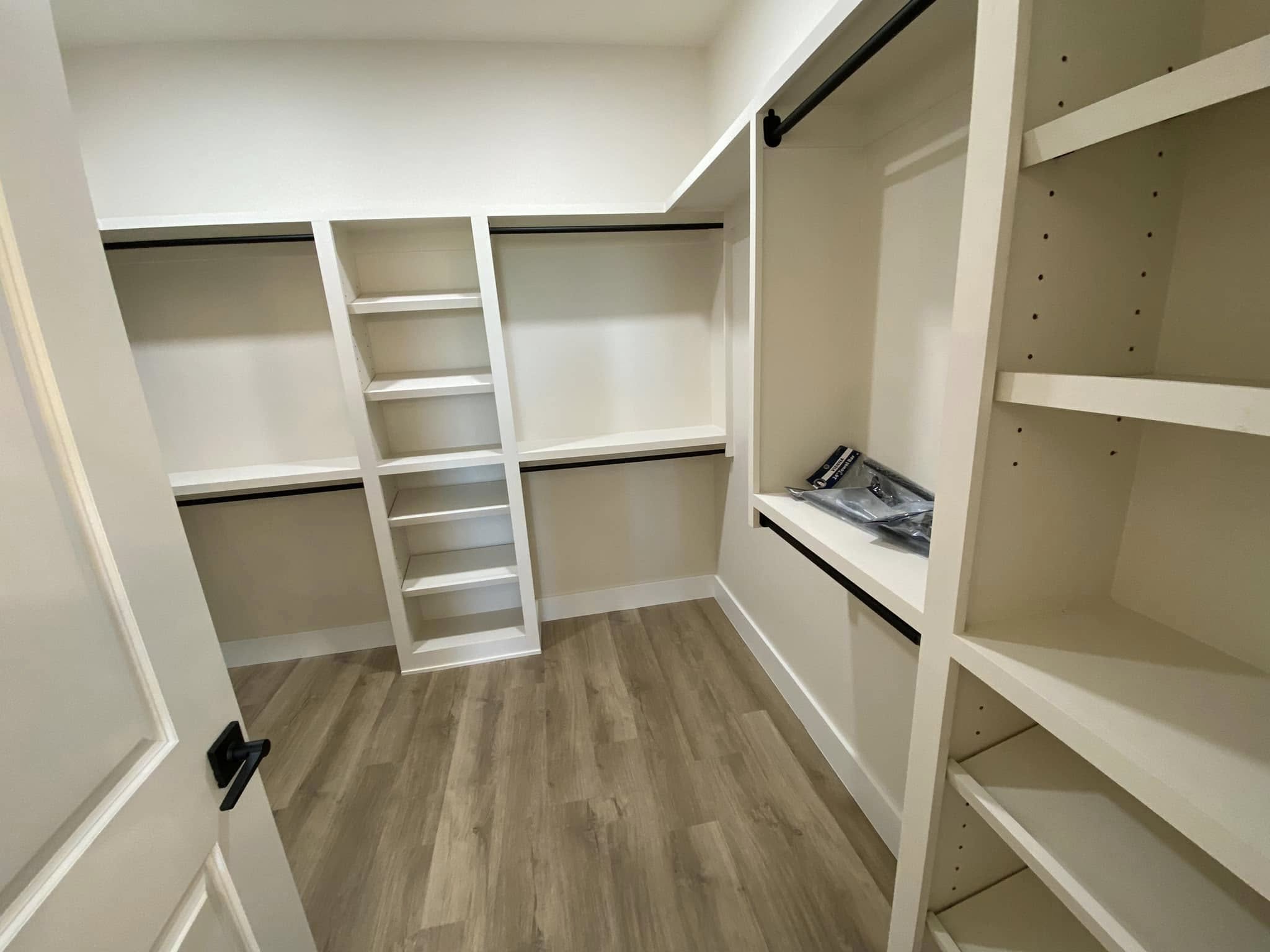 A spacious, empty walk-in closet with wooden flooring and white shelves. It has multiple hanging rods and open shelving for storage. The tone is organized and clean.