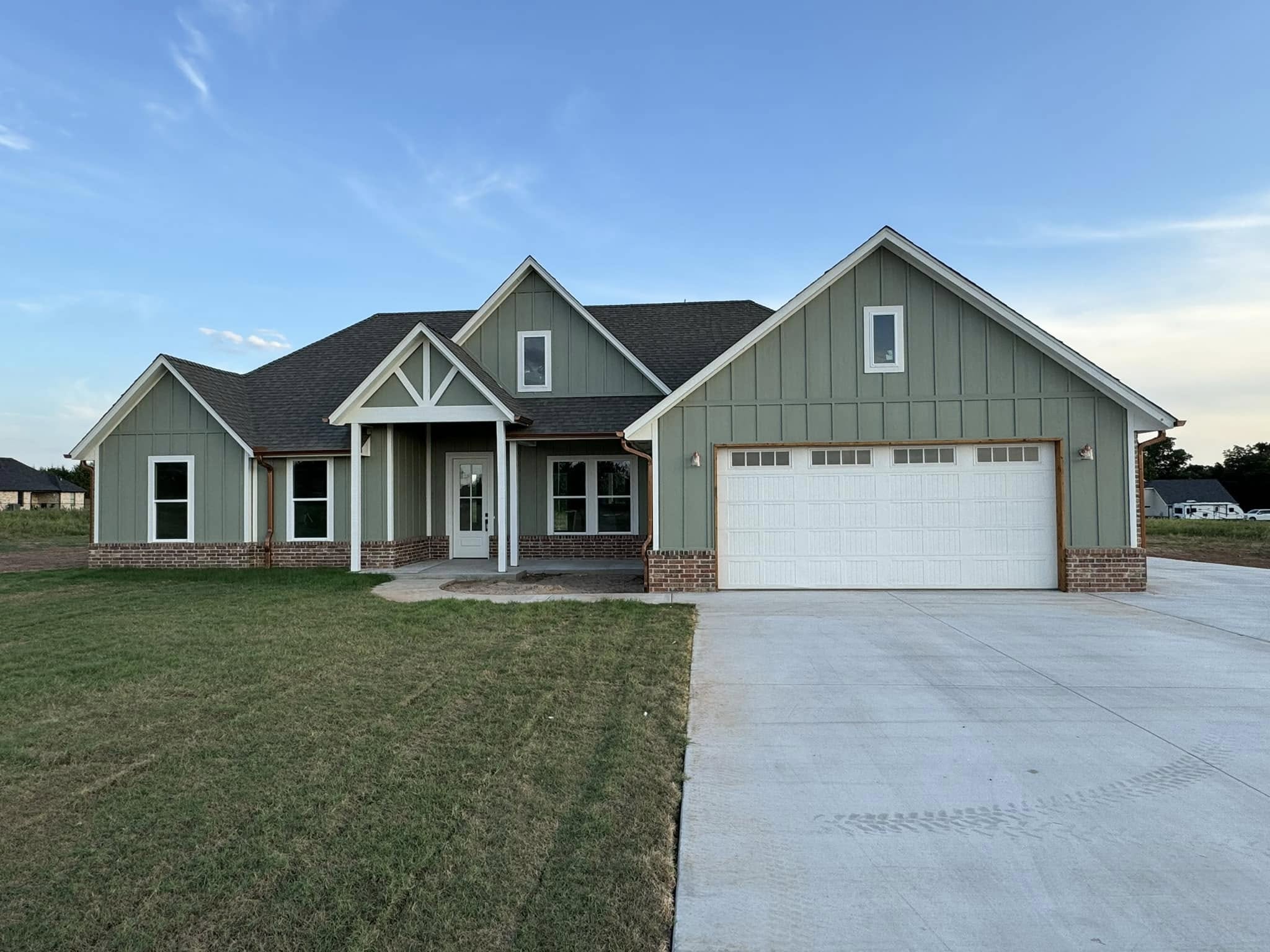 A modern single-story house with green and white siding, a large driveway, and a well-manicured lawn under a clear blue sky, conveying tranquility.