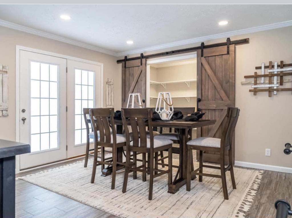Cozy dining room with a rustic table and six chairs, set on a patterned rug. Barn doors in the background reveal a shelved space. Bright with natural light.