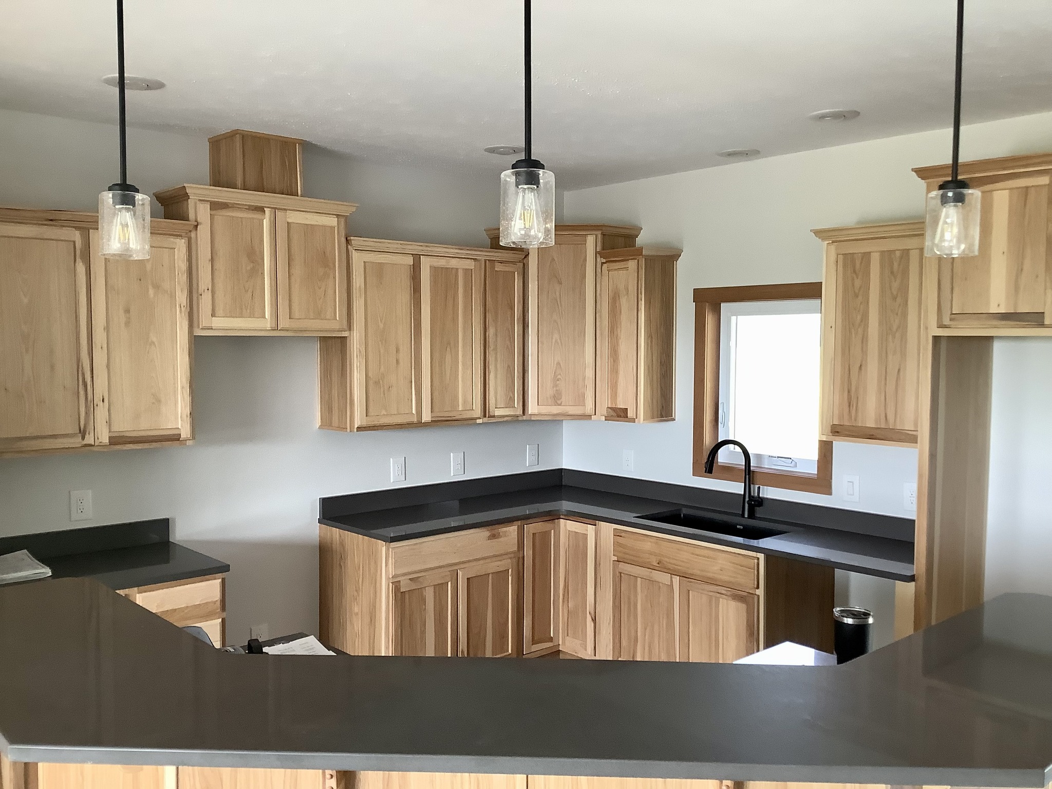 A modern kitchen with light wood cabinets, dark countertops, and black sink fixtures. Three pendant lights add warmth and style. Clean, orderly space.