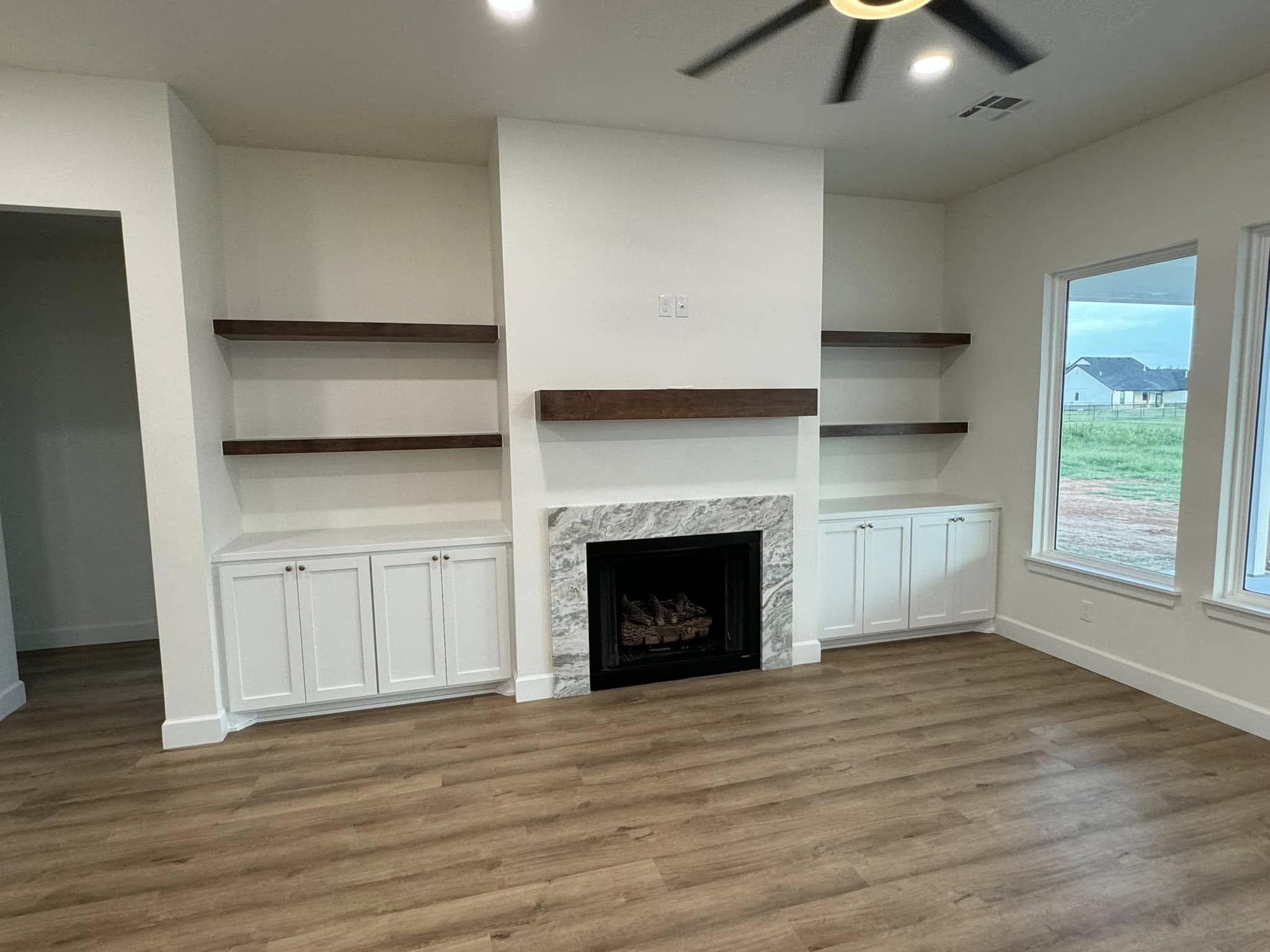 Modern living room with a central fireplace featuring a marble surround, flanked by white cabinets and dark wooden shelves. Large window on the right.