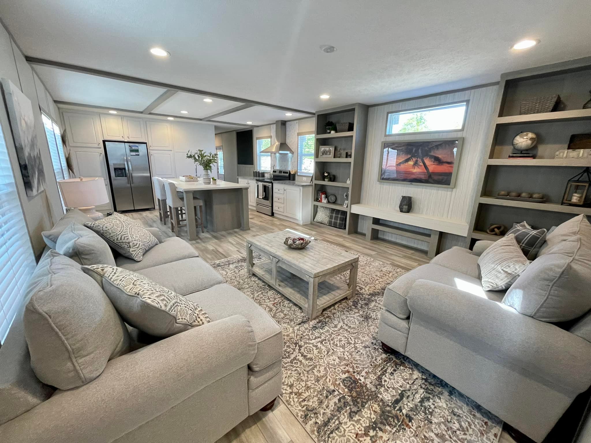 Modern living room and kitchen with a cozy feel. Neutral-toned sofas face each other across a wooden coffee table. Shelves decorate the wall. Kitchen in the background with island and dining chairs, creating an open and inviting space.